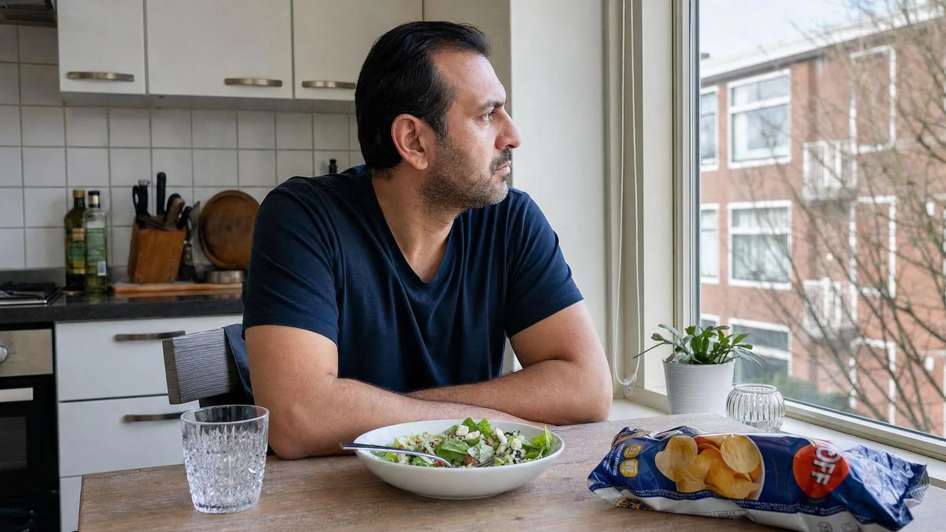 Iemand aan een keukentafel die naar buiten kijkt, met gezond eten en ongezonde snacks voor zich