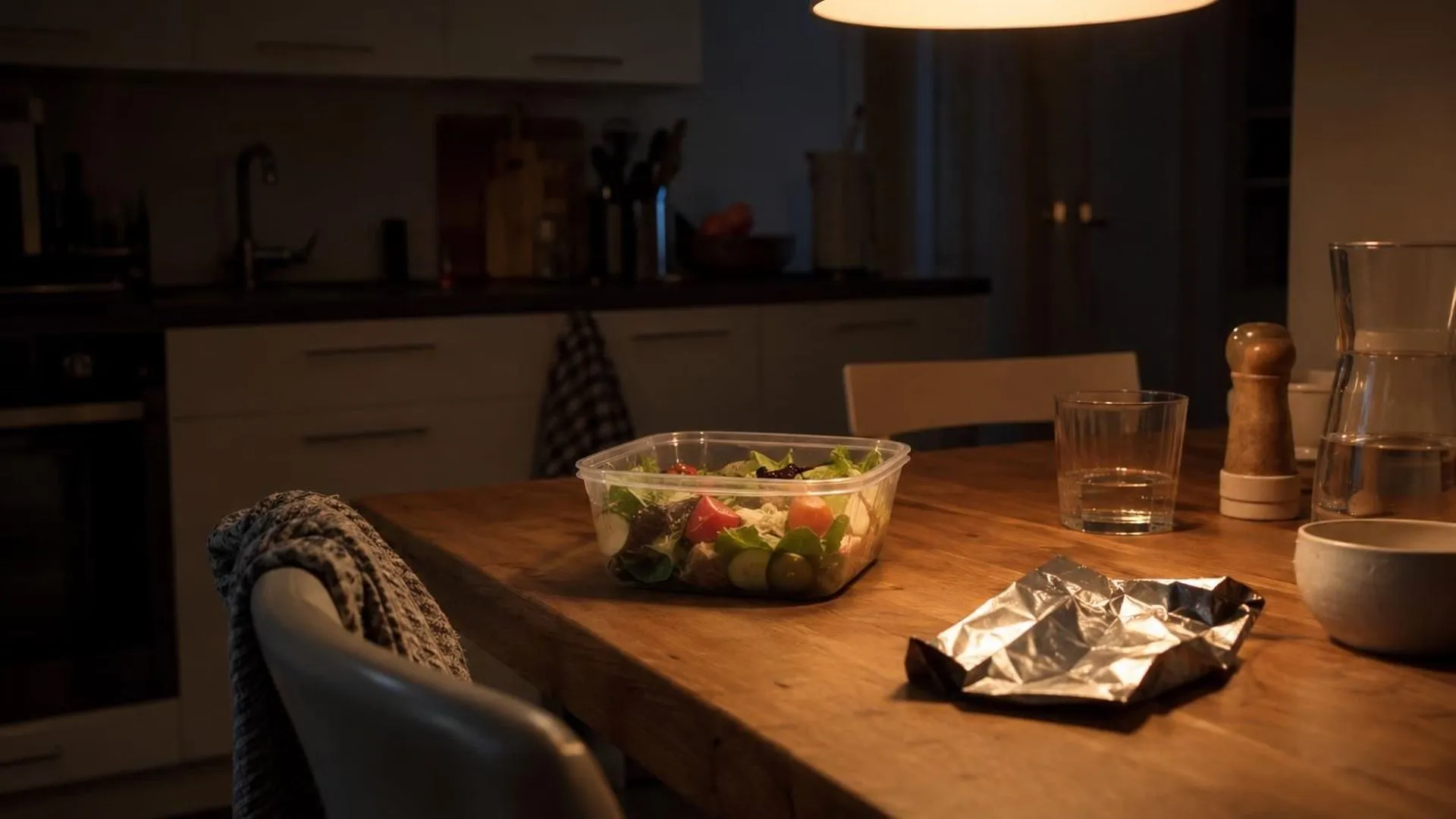 Onaangeroerde salade naast een leeg chocoladewikkel op een keukentafel in lamplicht