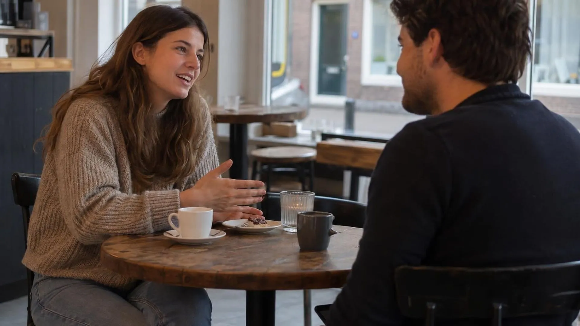 Twee mensen aan een cafétafel, de een praat open, de ander knikt maar houdt een telefoon net onder de tafelrand vast.