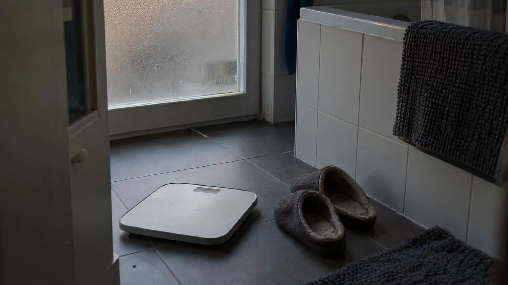 Een weegschaal op de badkamervloer naast versleten pantoffels in schemerlicht.