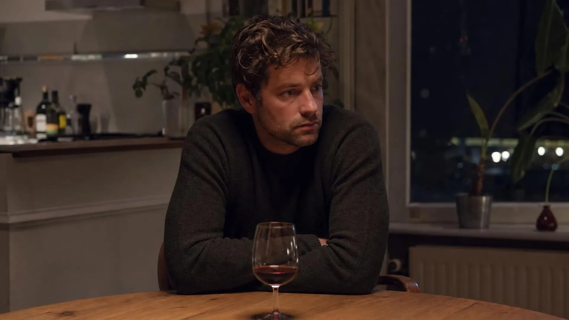 Persoon aan keukentafel 's avonds met glas wijn, in gedachten verzonken