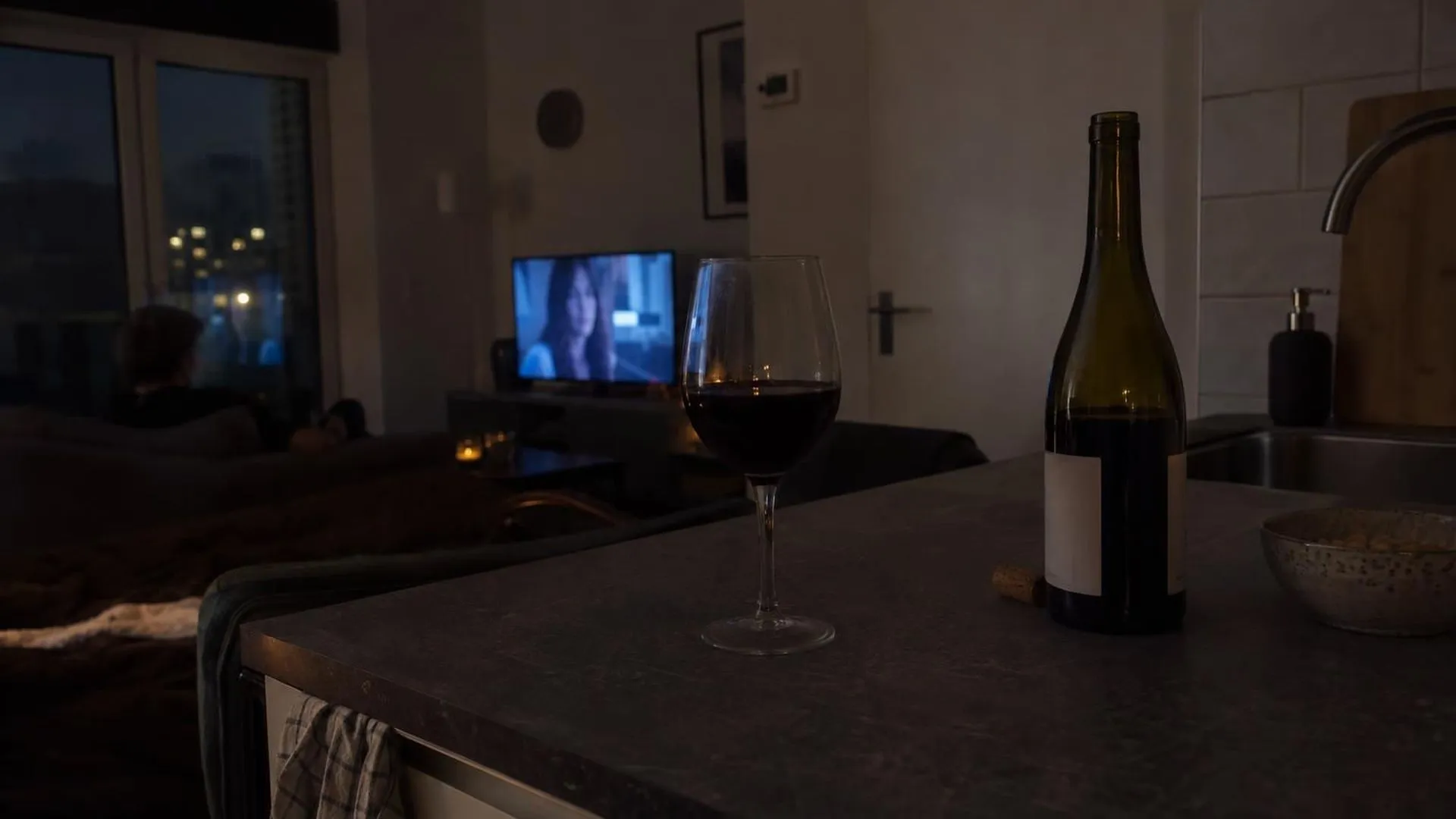 Een wijnglas en geopende fles op het aanrecht in de avond, met het schijnsel van een televisie op de achtergrond