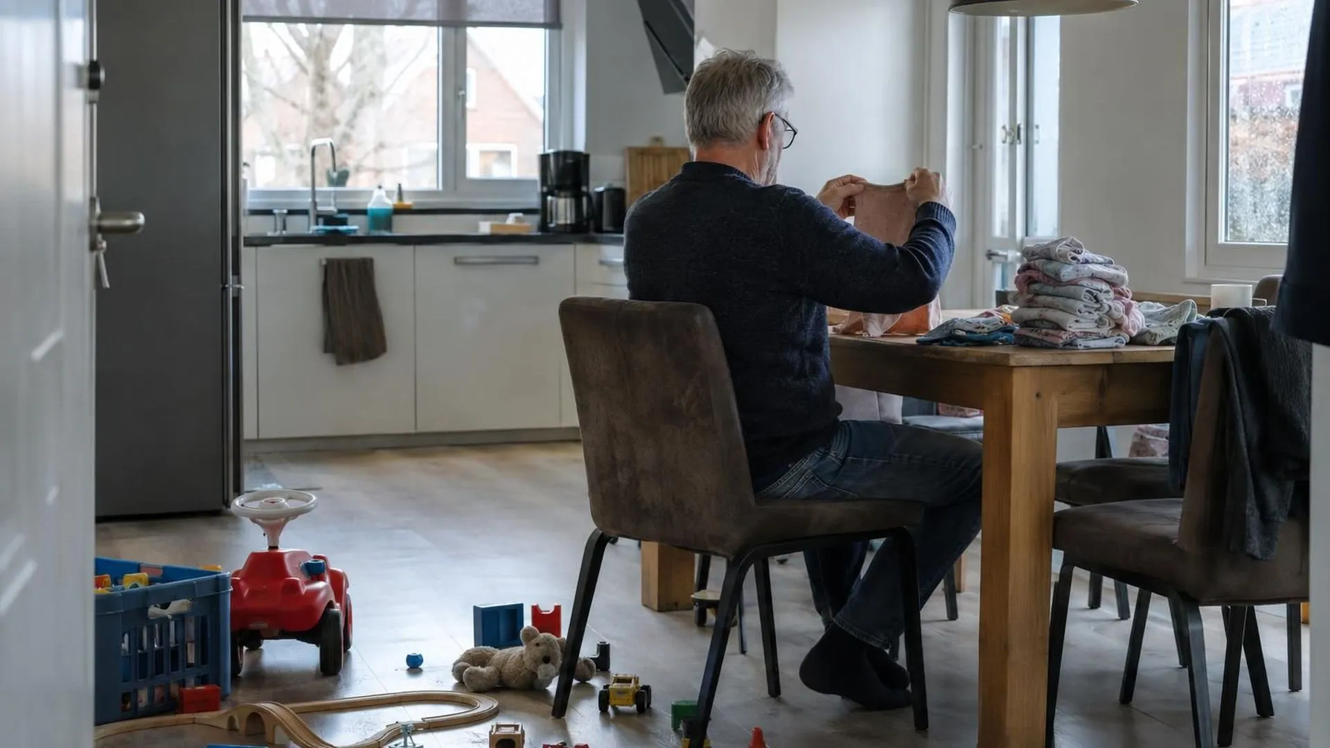 Iemand van achteren gezien, vouwt kinderkleren aan de keukentafel op een vroege zondagochtend, speelgoed op de vloer