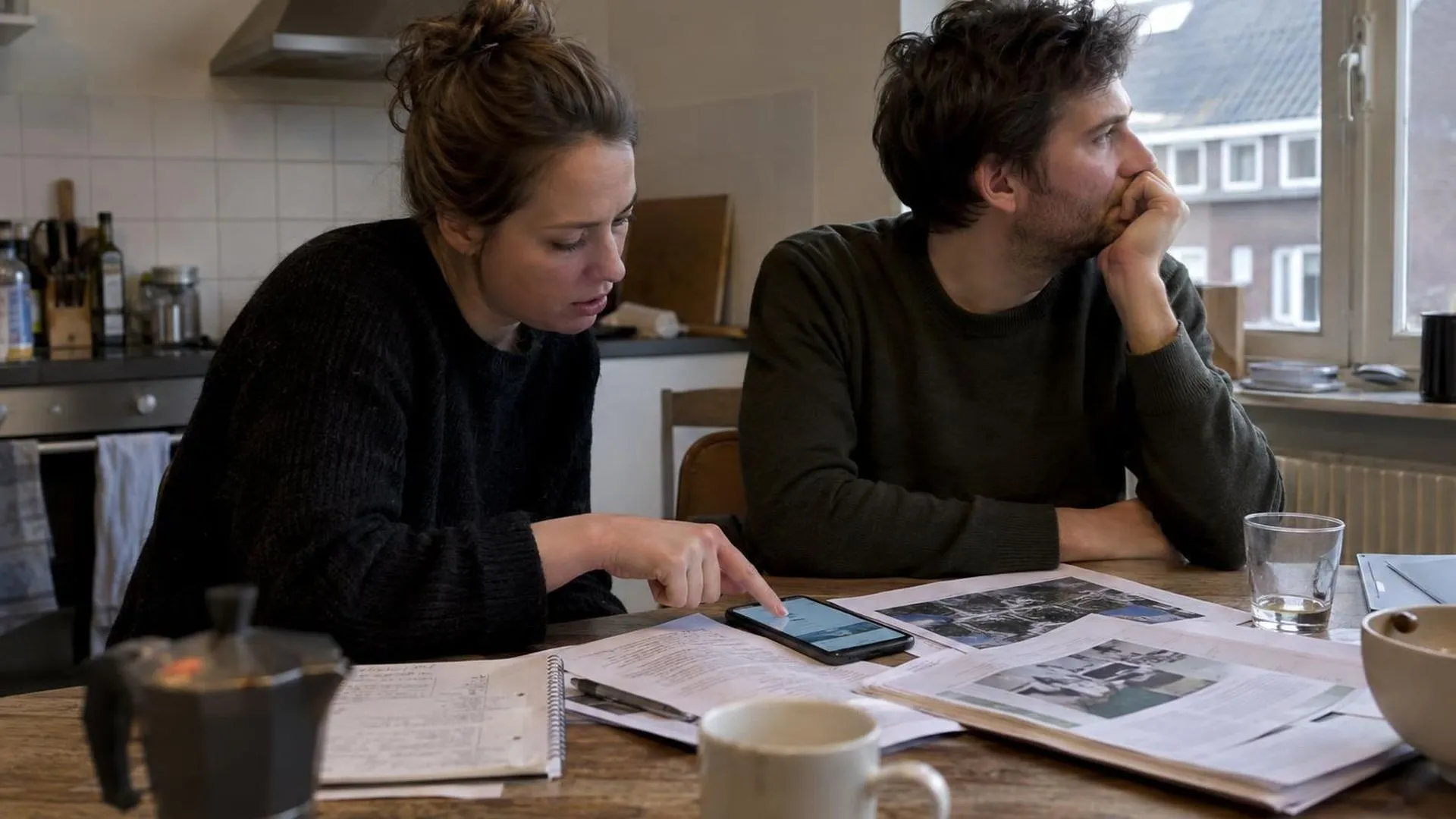 Twee mensen aan een keukentafel, de een wijst dringend naar een telefoonscherm vol artikelen, de ander kijkt weg.