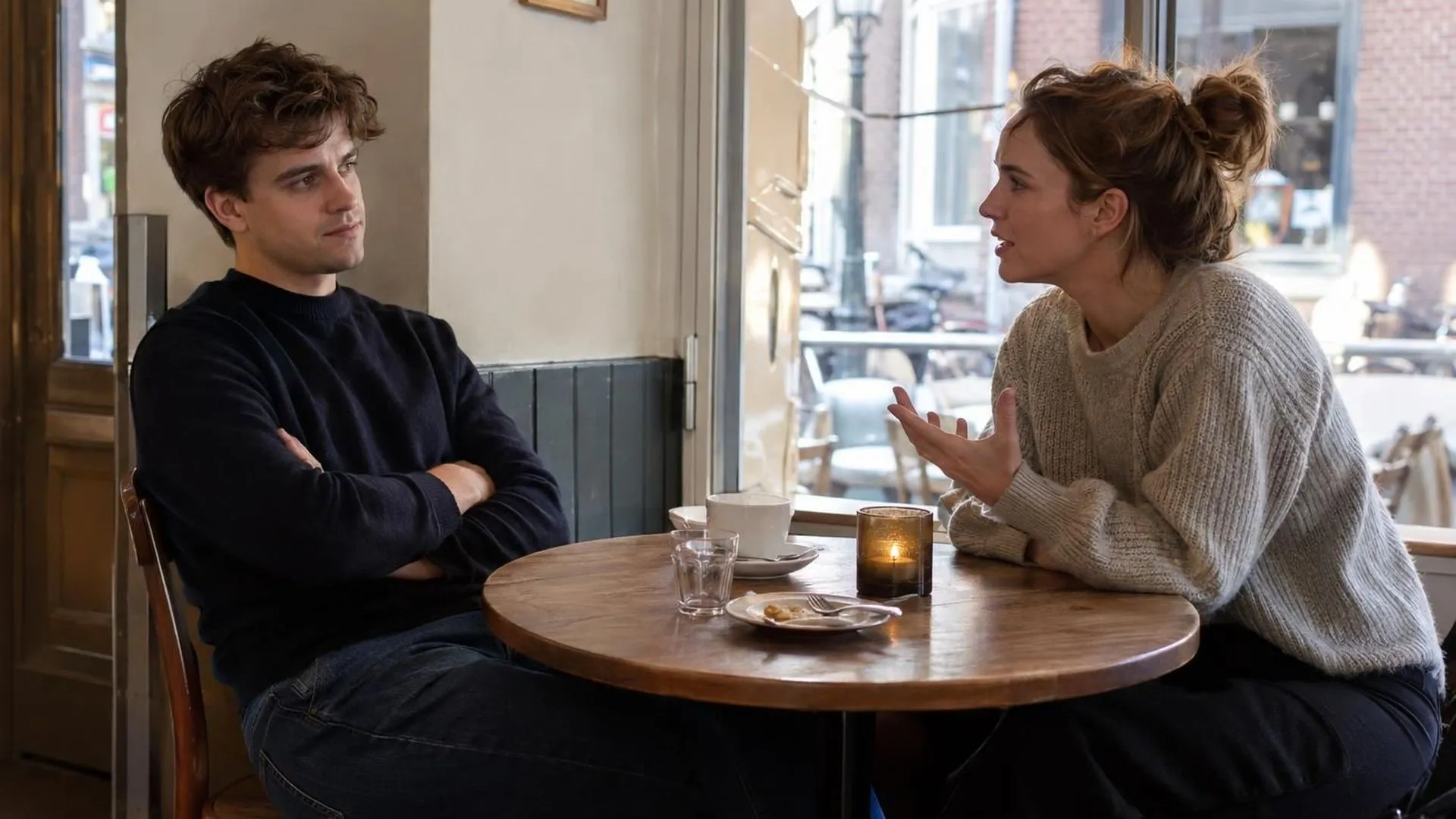Twee mensen aan een cafetafel, de een met over elkaar geslagen armen, de ander leunt naar voren