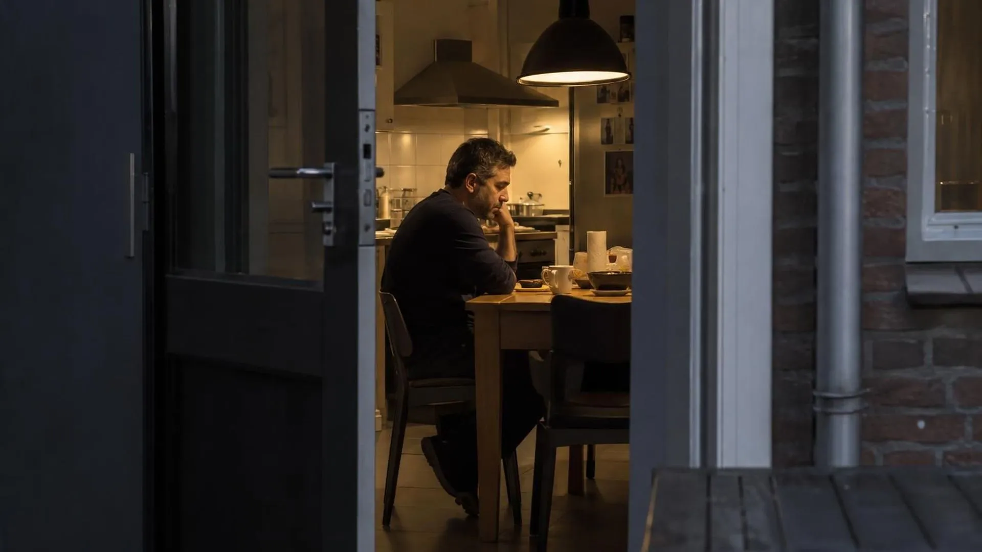 Door een op een kier staande deur gezien: iemand zit zwijgend aan de keukentafel.