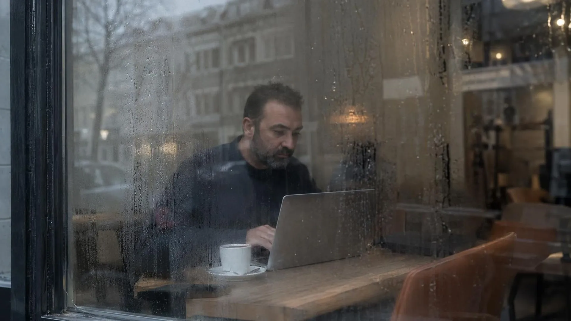 Door een beslagen caféraam gezien zit iemand binnen achter een laptop, het glas vervaagt het beeld