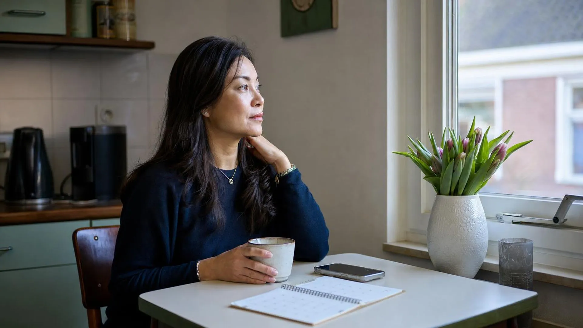 Mantelzorger zit aan tafel en kijkt vermoeid uit het raam