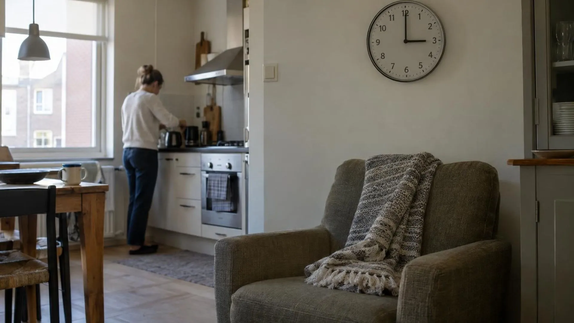 Een keukenklok wijst 15.00 uur aan naast een lege fauteuil met een opgevouwen plaid