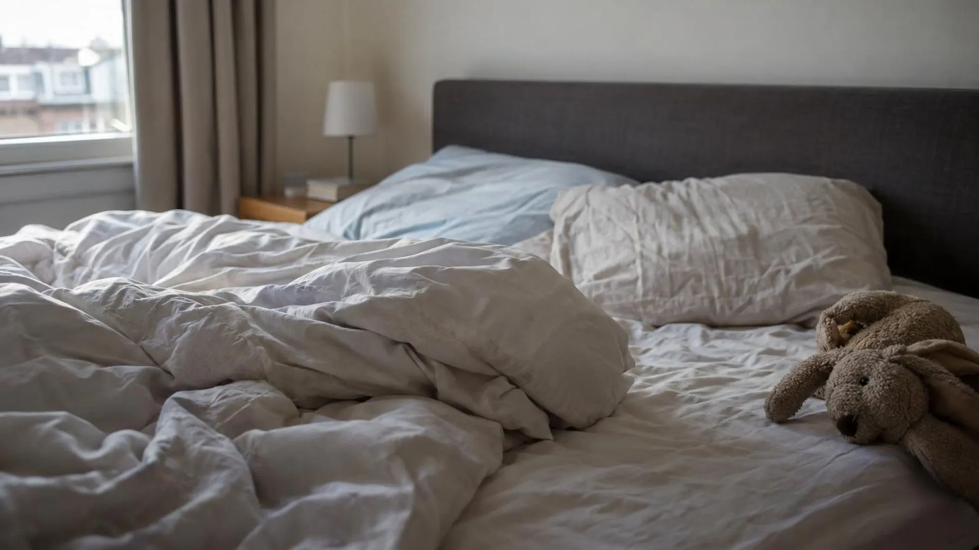Close-up van verkreukelde lakens in het ochtendlicht van een zaterdag, met een knuffel van een kind aan de rand van het bed.