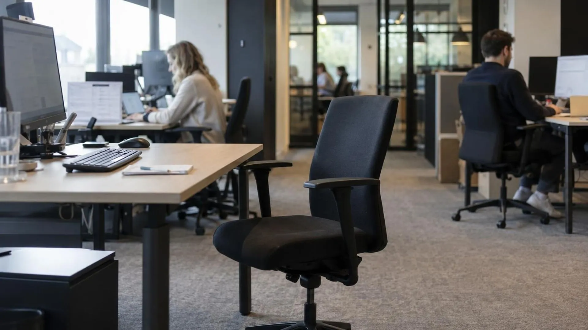 Een lege bureaustoel, teruggerold van het bureau, in een kantoor met wazige collega's op de achtergrond