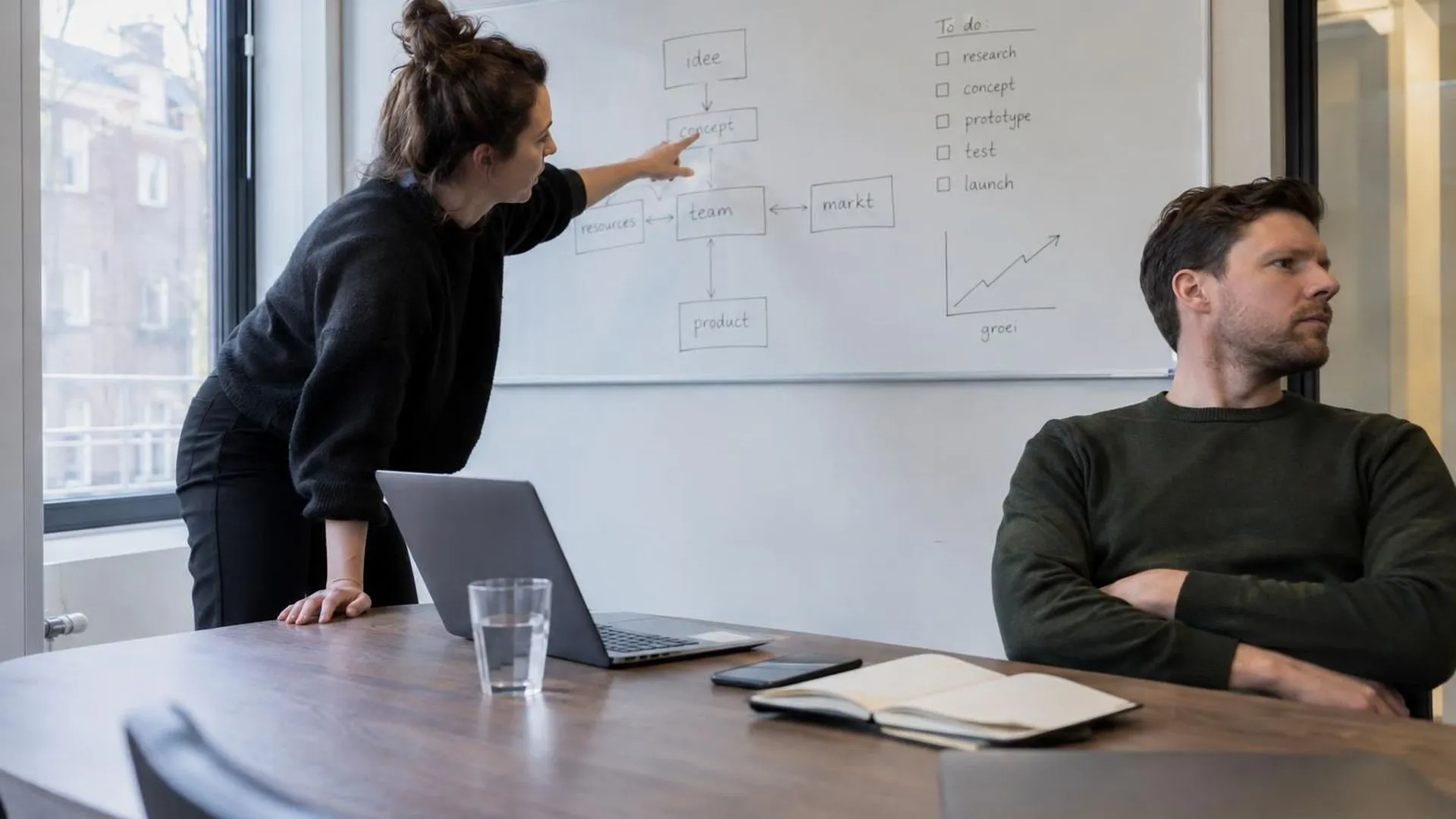 Twee collega's in een vergadering, de een leunt naar voren en wijst, de ander leunt achterover met over elkaar geslagen armen