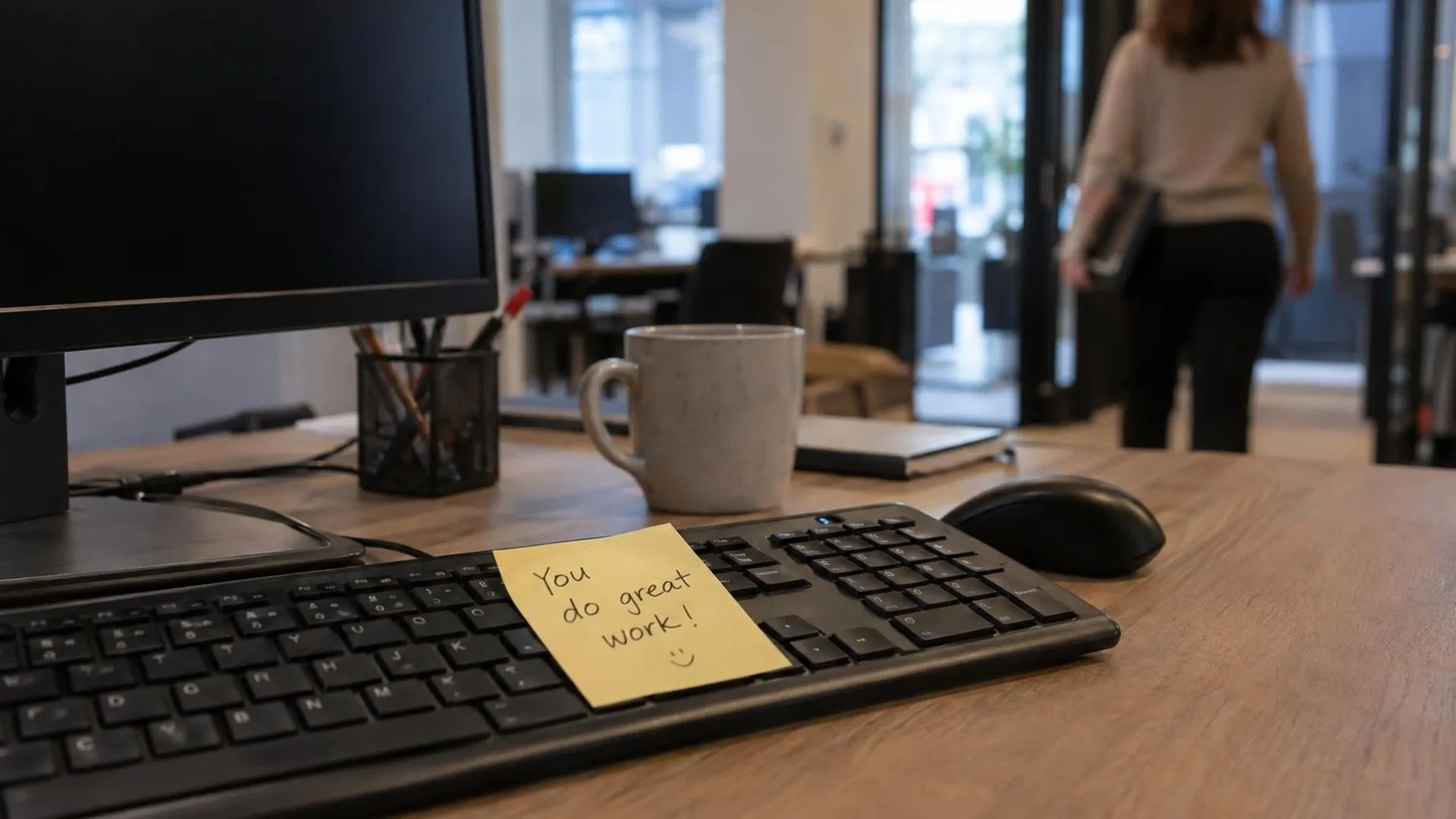 Een geel Post-it-briefje met een compliment op een leeg toetsenbord, een collega loopt wazig op de achtergrond weg.