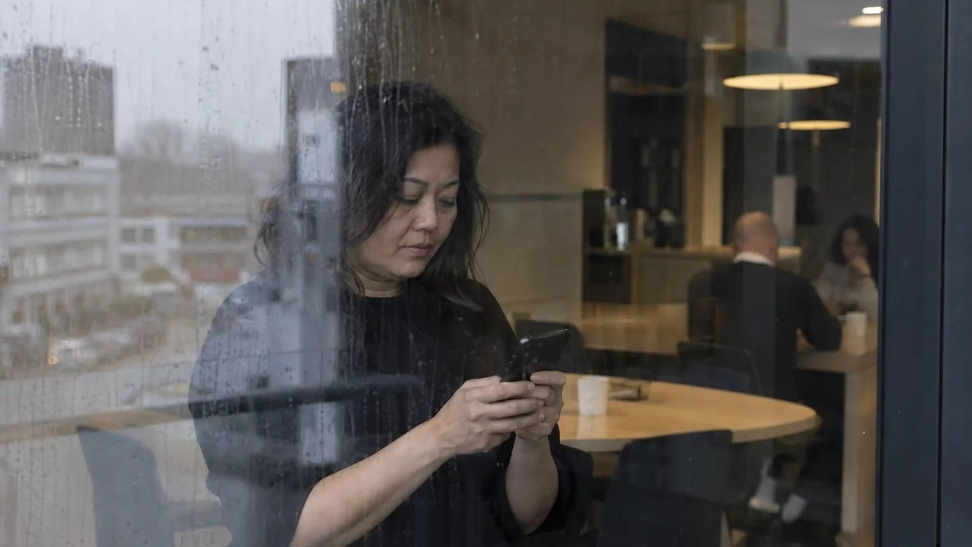 Door een beregend kantoorraam gezien: iemand die tijdens de pauze afleiding zoekt op de telefoon