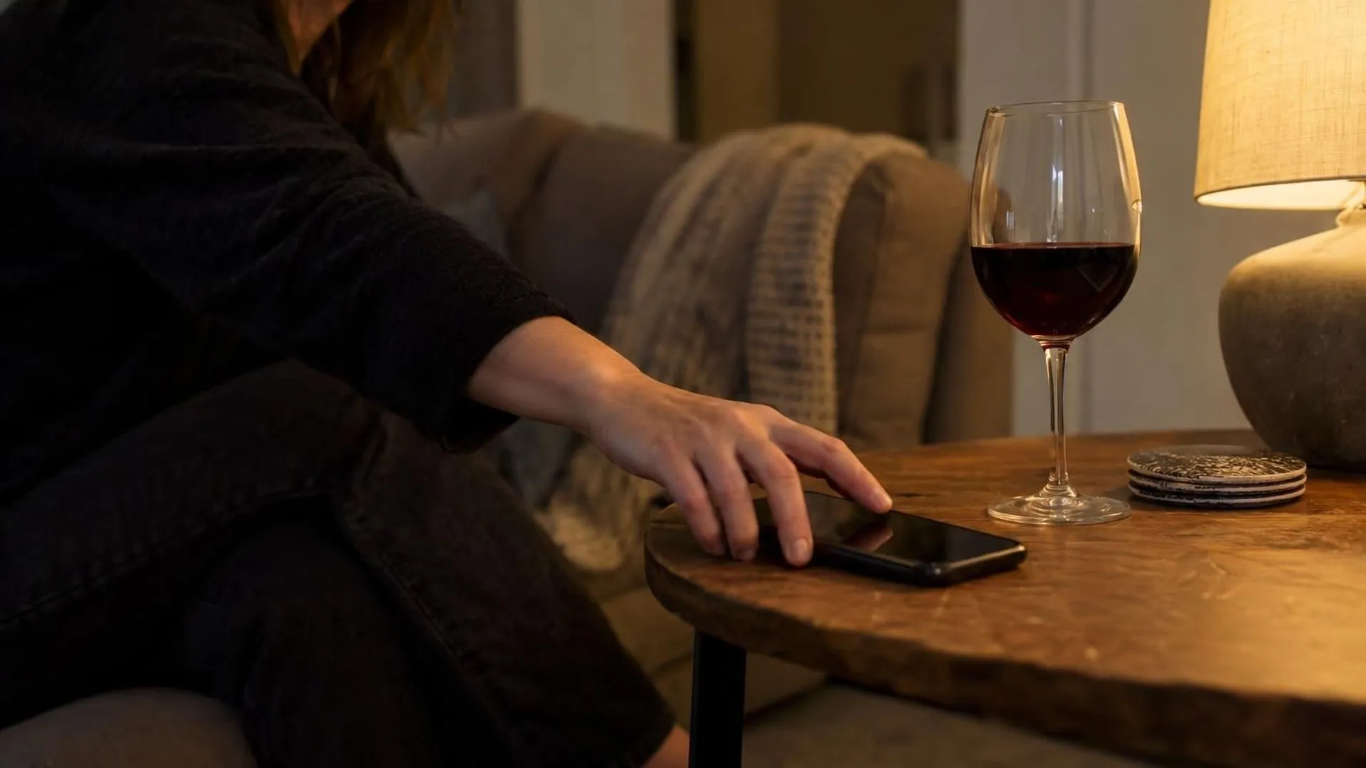 Een hand reikt naar een telefoon op een bijzettafel naast een onaangeroerd glas wijn, bij warm avondlicht.
