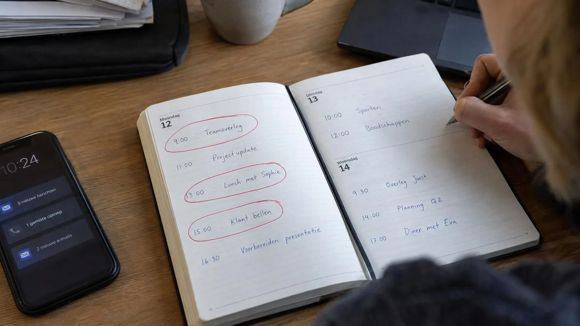 Een opengeslagen bureau-agenda met meerdere taken in rode inkt omcirkeld op dezelfde dag, telefoon trilt ernaast.