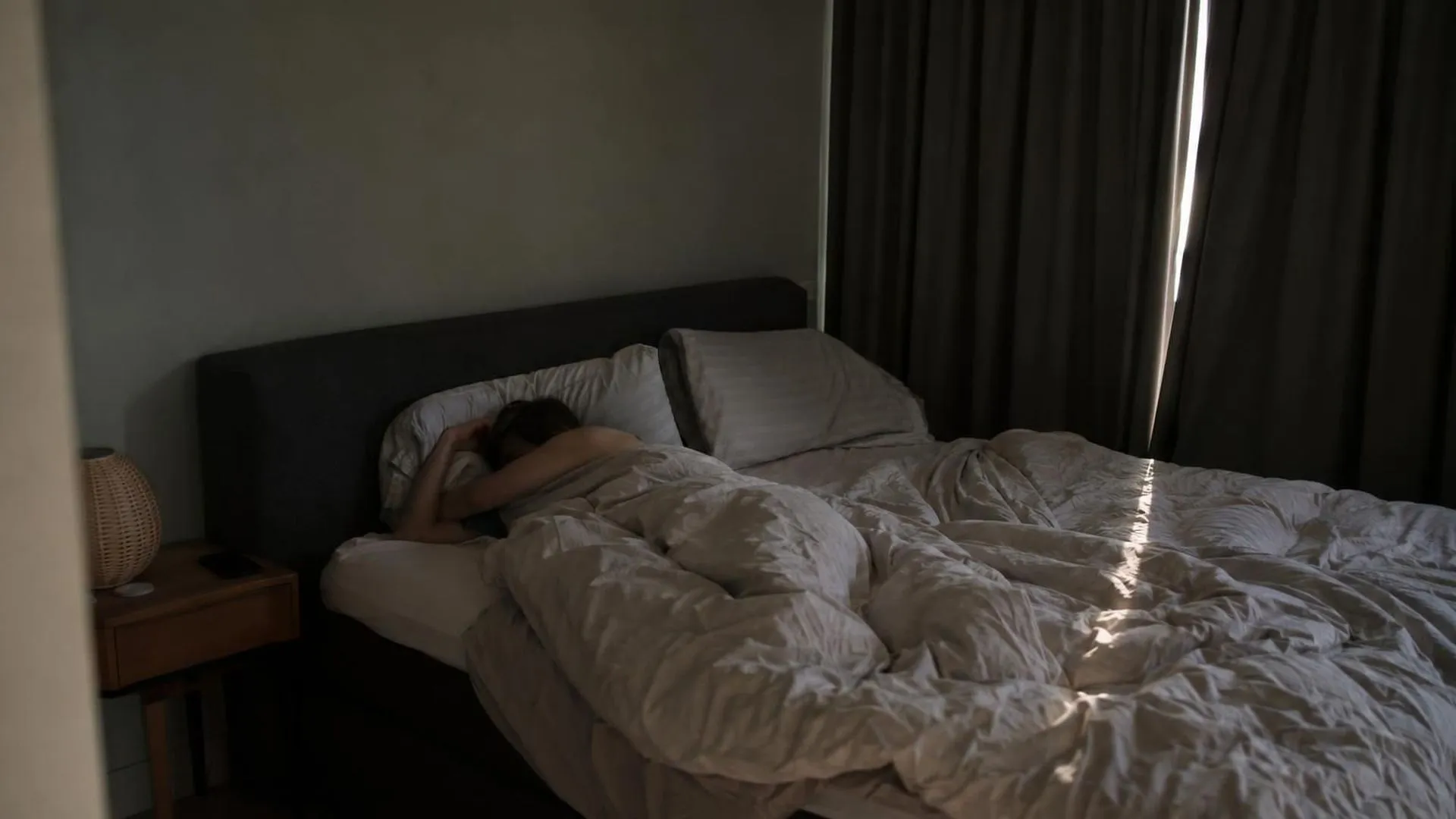 Een onopgemaakt bed op klaarlichte dag met dichte gordijnen en een smalle lichtstreep over de lakens