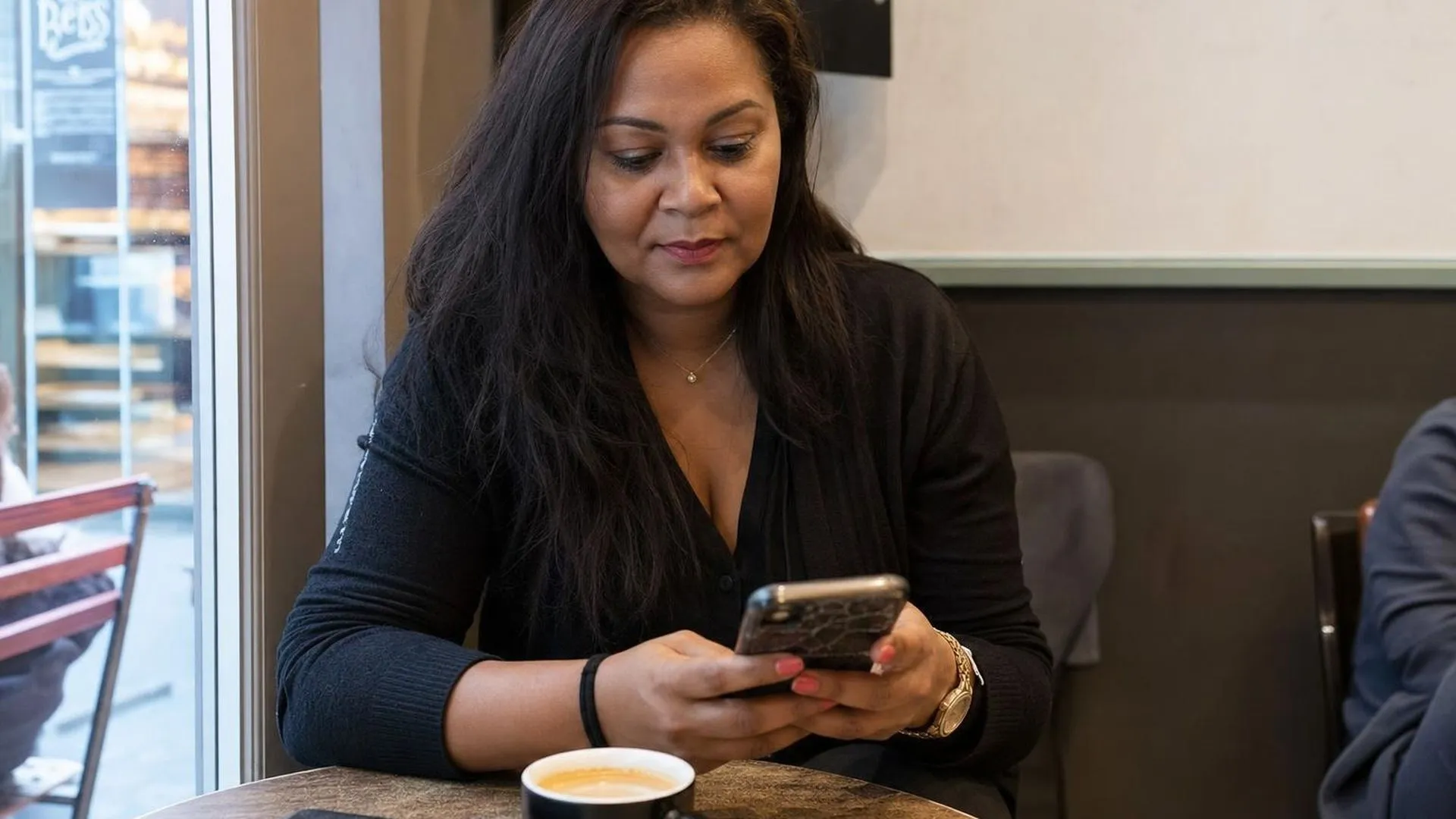 Persoon zit aan een cafétafel met telefoon in hand en een onaangeroerd kopje koffie
