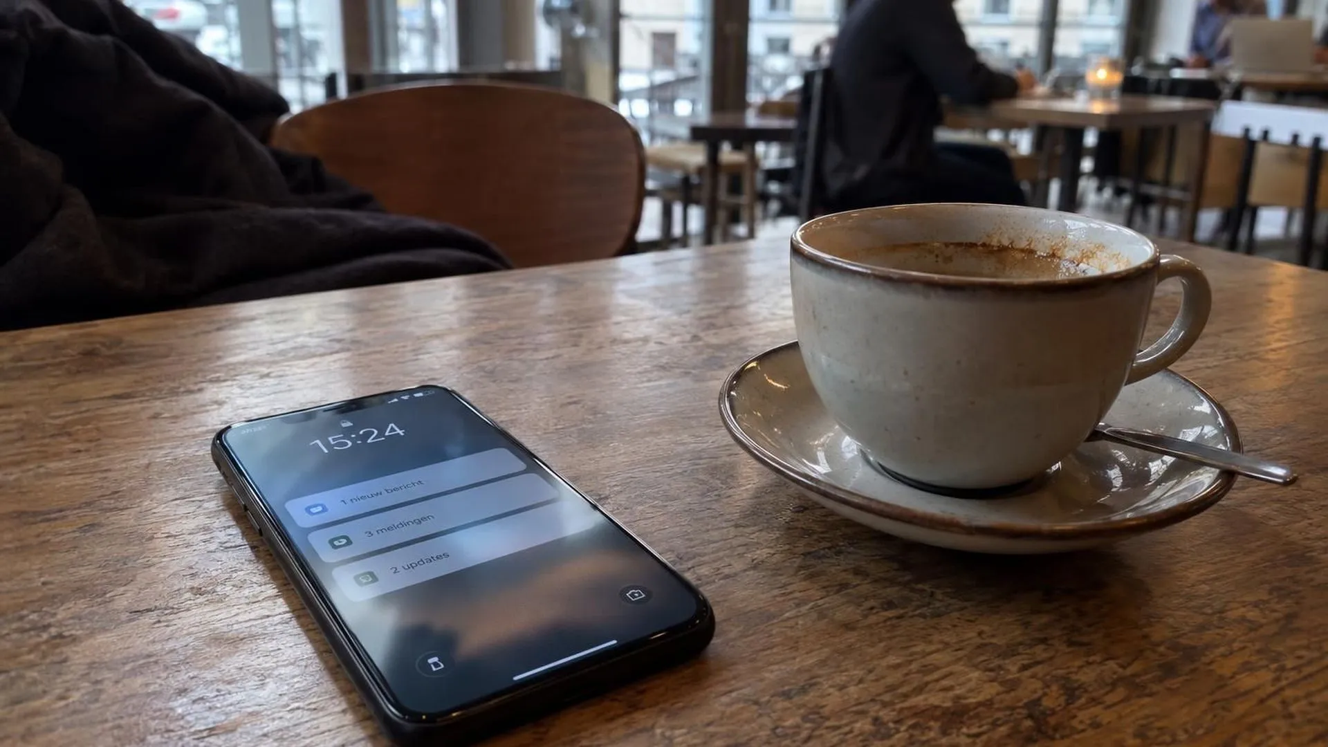 Een telefoon met oplichtend scherm op een cafetafel naast een onaangeroerd, koud wordend kopje koffie.