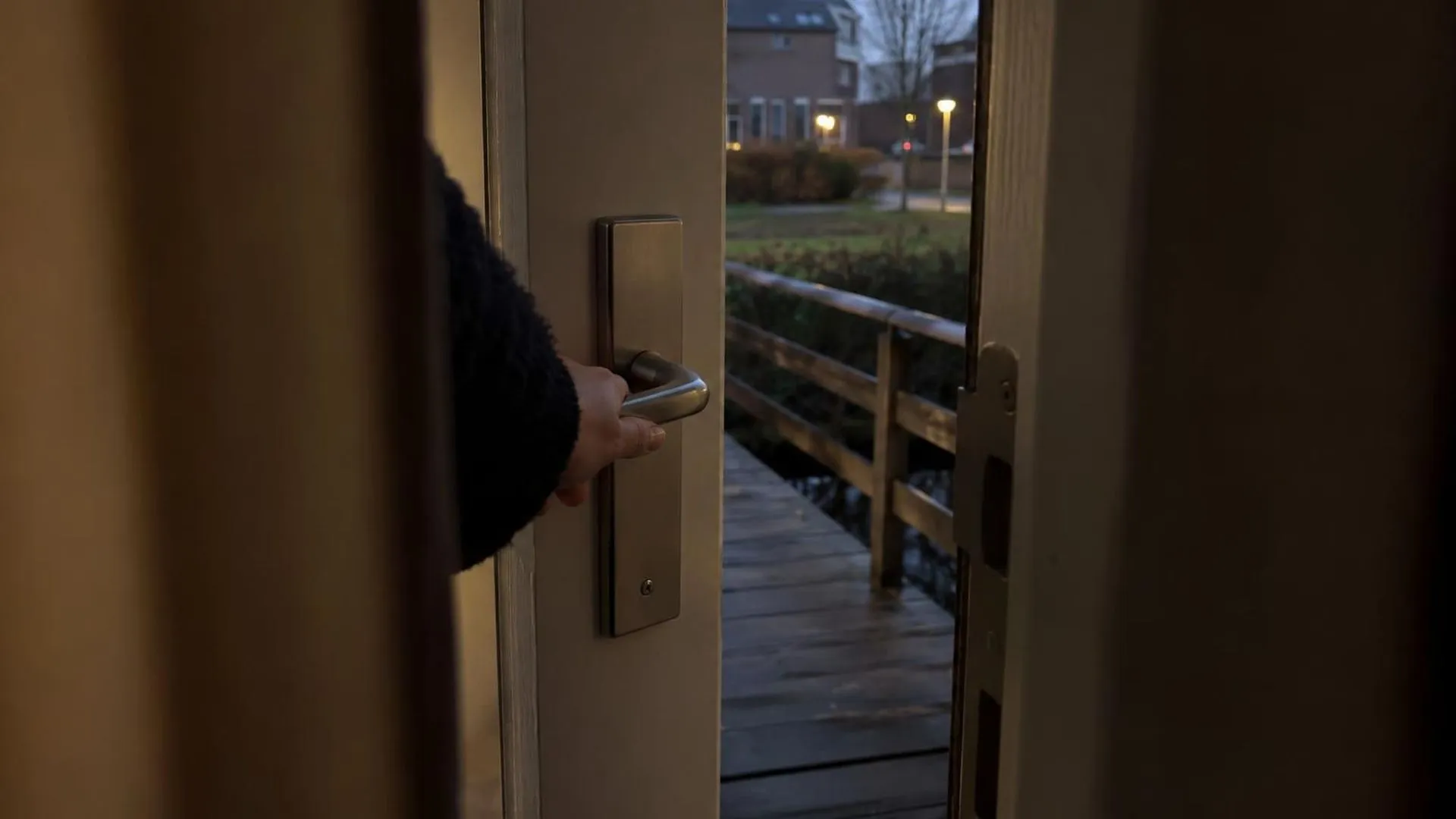 Een hand rust op een deurklink, gezien door de kier van een op een kiertje staande voordeur, bij schemerlicht.