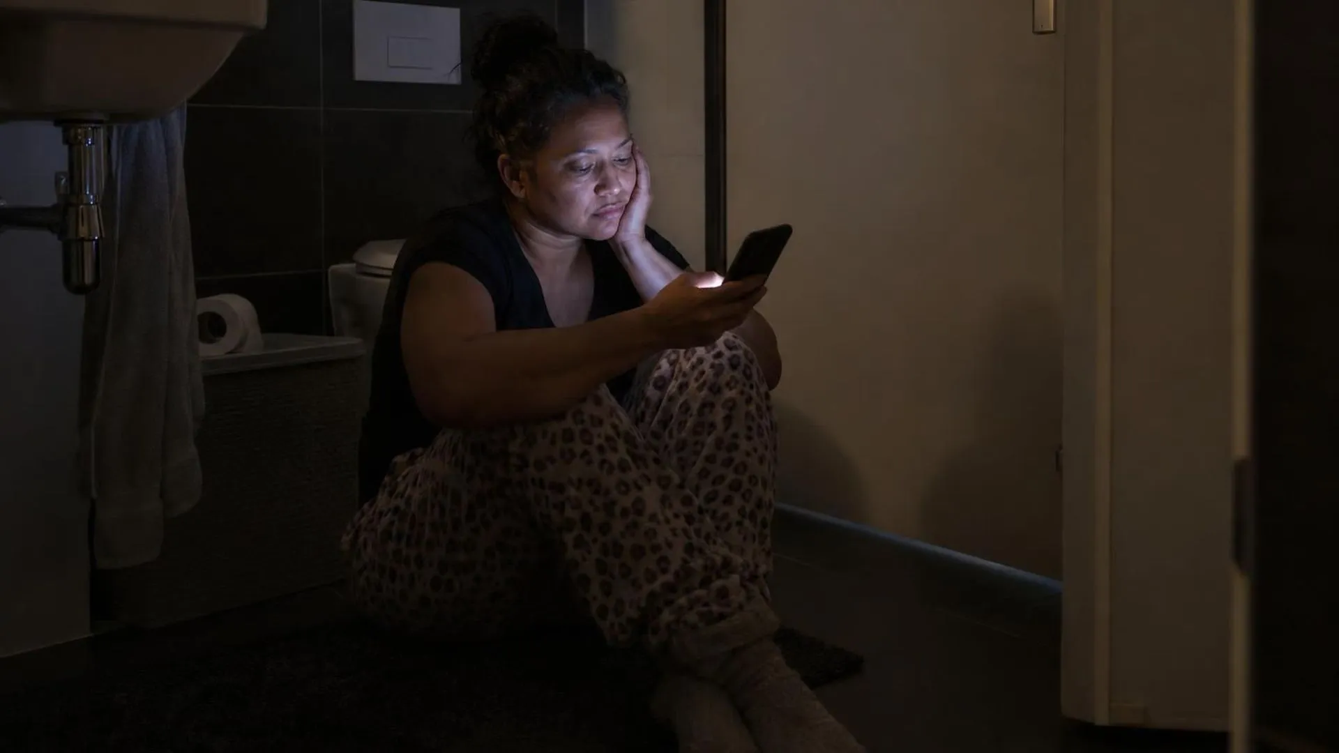 Iemand zit 's nachts op de badkamervloer met de deur dicht, alleen het licht van een telefoon zichtbaar