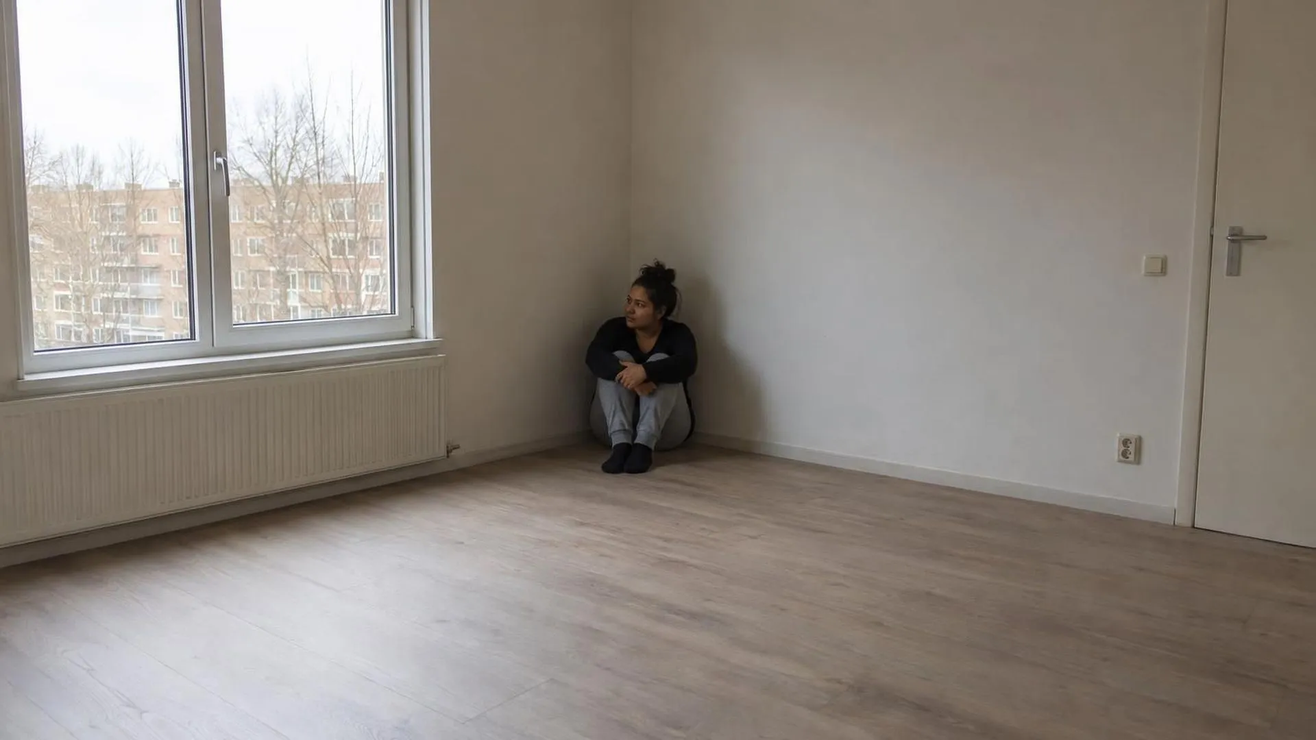 Een brede opname toont iemand klein in de hoek van een kale, grote kamer, met veel lege ruimte eromheen.