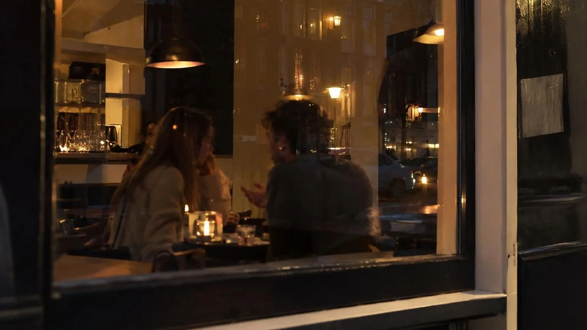 Door een caferaam gezien 's avonds, met twee wazige silhouetten in gesprek binnen en straatlicht dat weerspiegelt in het glas
