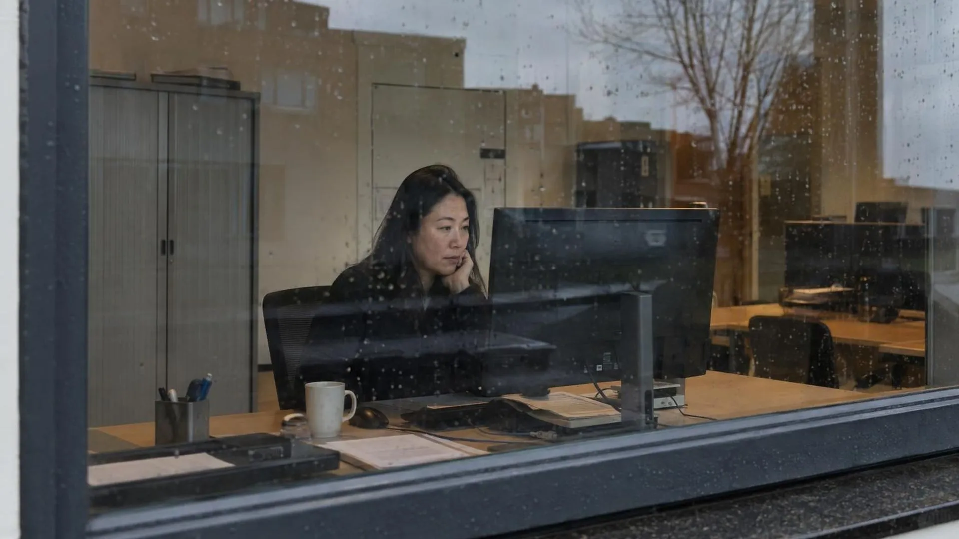Door een beregend raam gezien: iemand zit roerloos achter een bureau terwijl reflecties over het glas vallen.