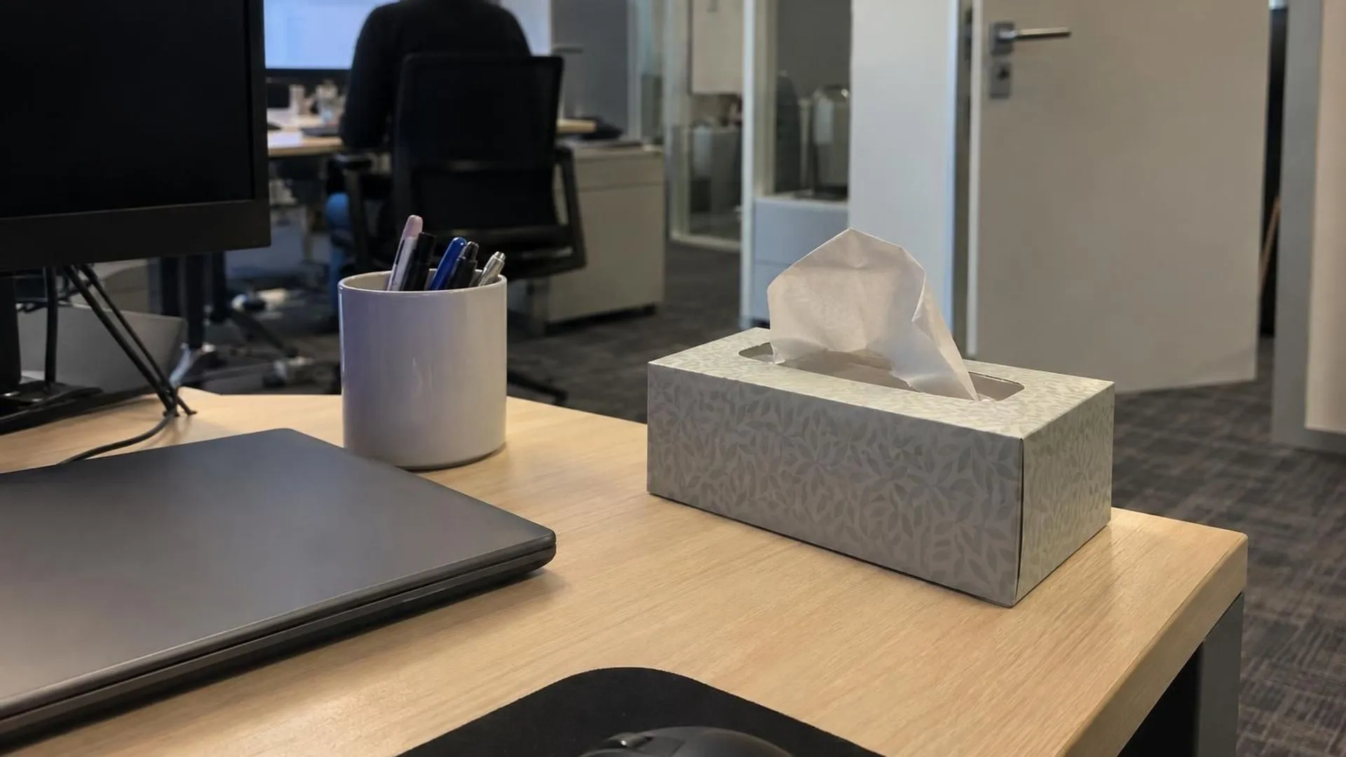 Een half leeg doosje tissues staat op de hoek van een bureau naast een dichte laptop, op een gewone kantoorochtend.