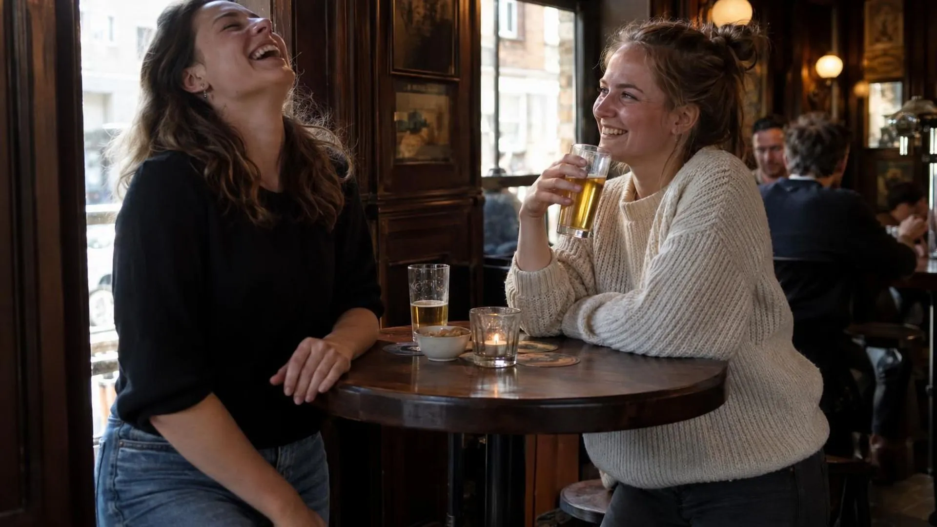 Twee mensen staan aan een statafel in een bruin café, de een lacht met het hoofd achterover, de ander glimlacht met een biertje in de hand.