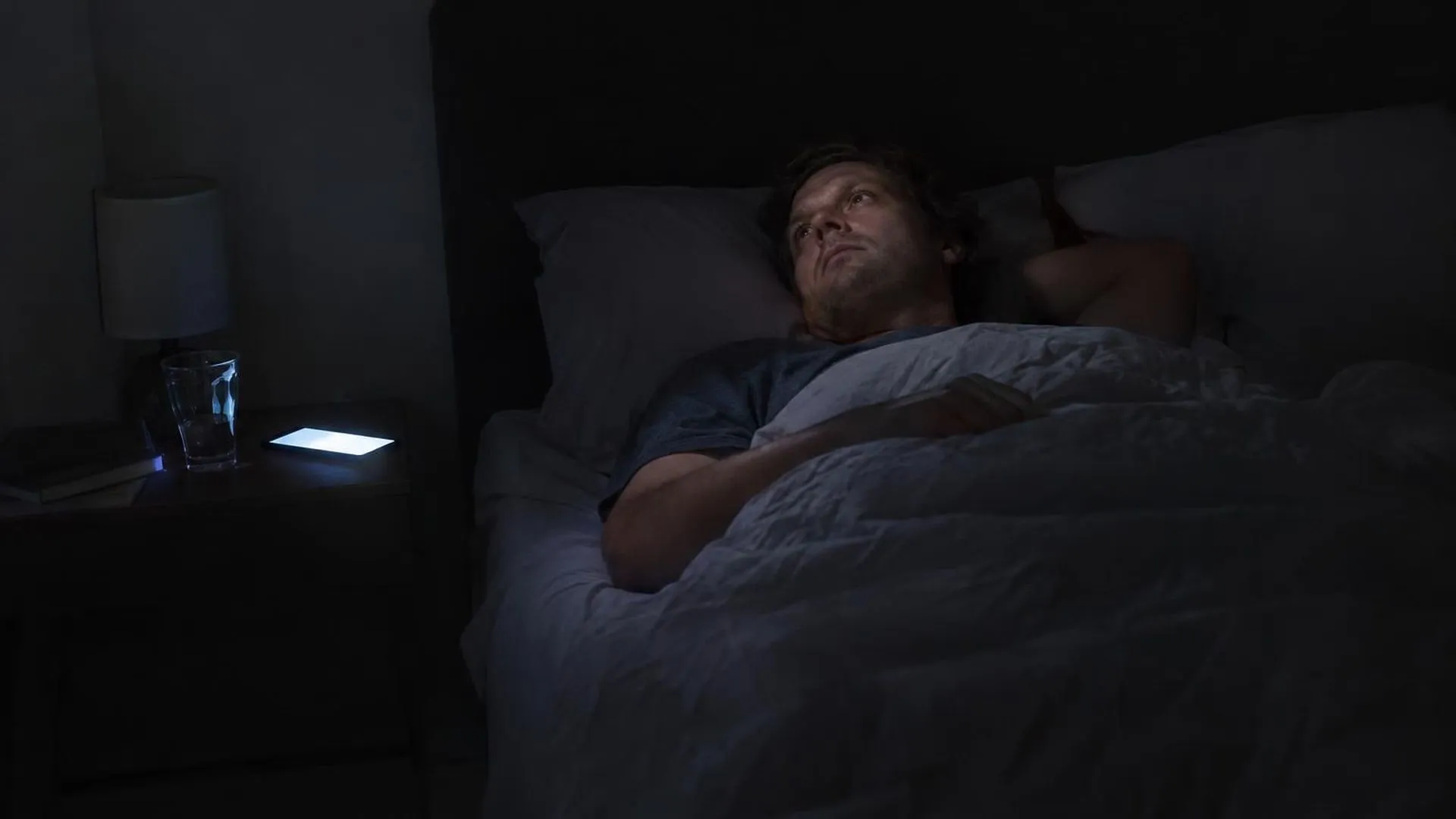 Iemand ligt 's nachts met open ogen in bed, alleen het scherm van een telefoon op het nachtkastje verlicht de verder donkere kamer.