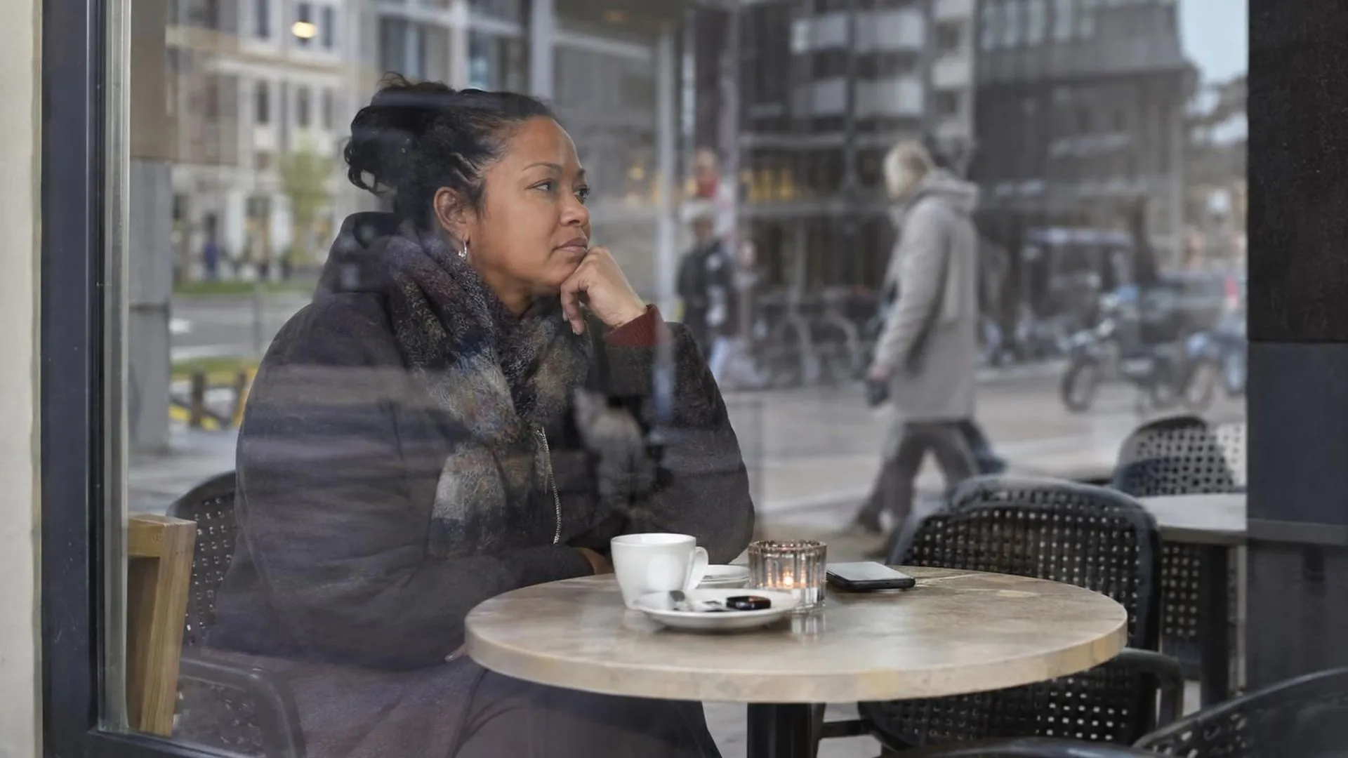 Door een caféraam heen zit iemand alleen aan een terrastafel, met vage reflecties van voorbijgangers over het glas.