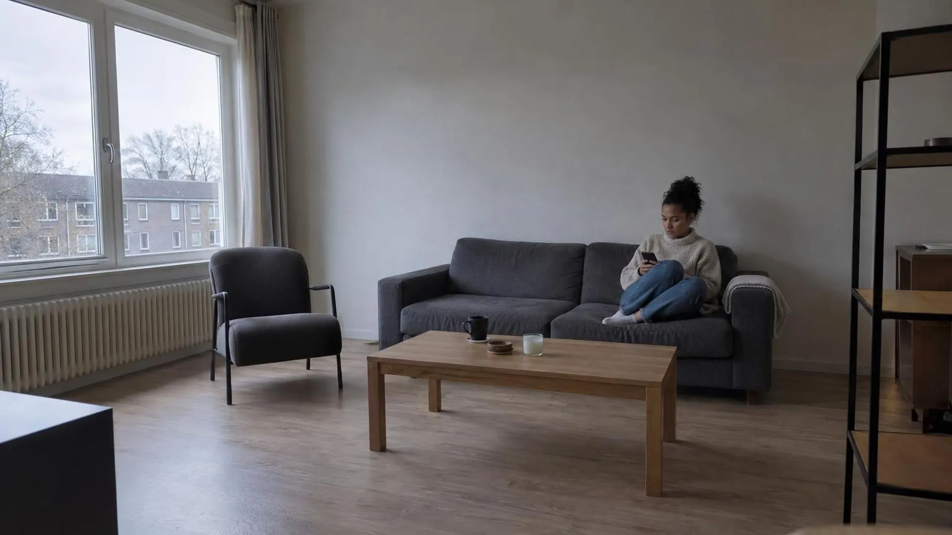 Iemand zit klein op de bank in een sober ingerichte huiskamer bij vlak grijs daglicht.
