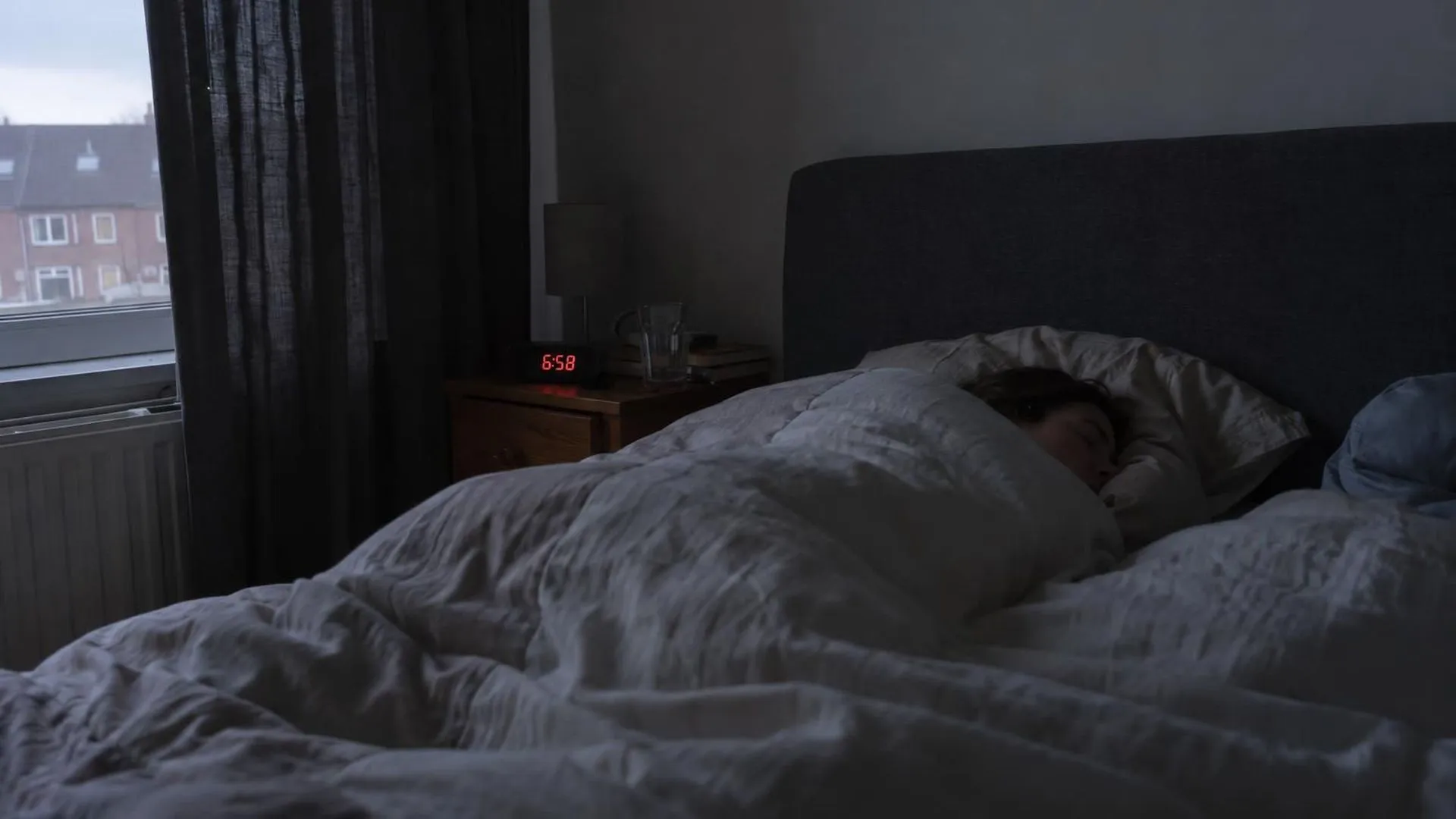 Een slaapkamer vlak voor zonsopgang, een wekker geeft 6:58 aan en de dekens liggen nog hoog opgetrokken