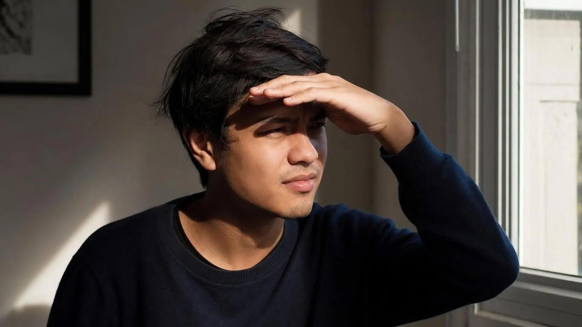 Iemand schermt de ogen af tegen fel licht bij een raam in een rustige kamer