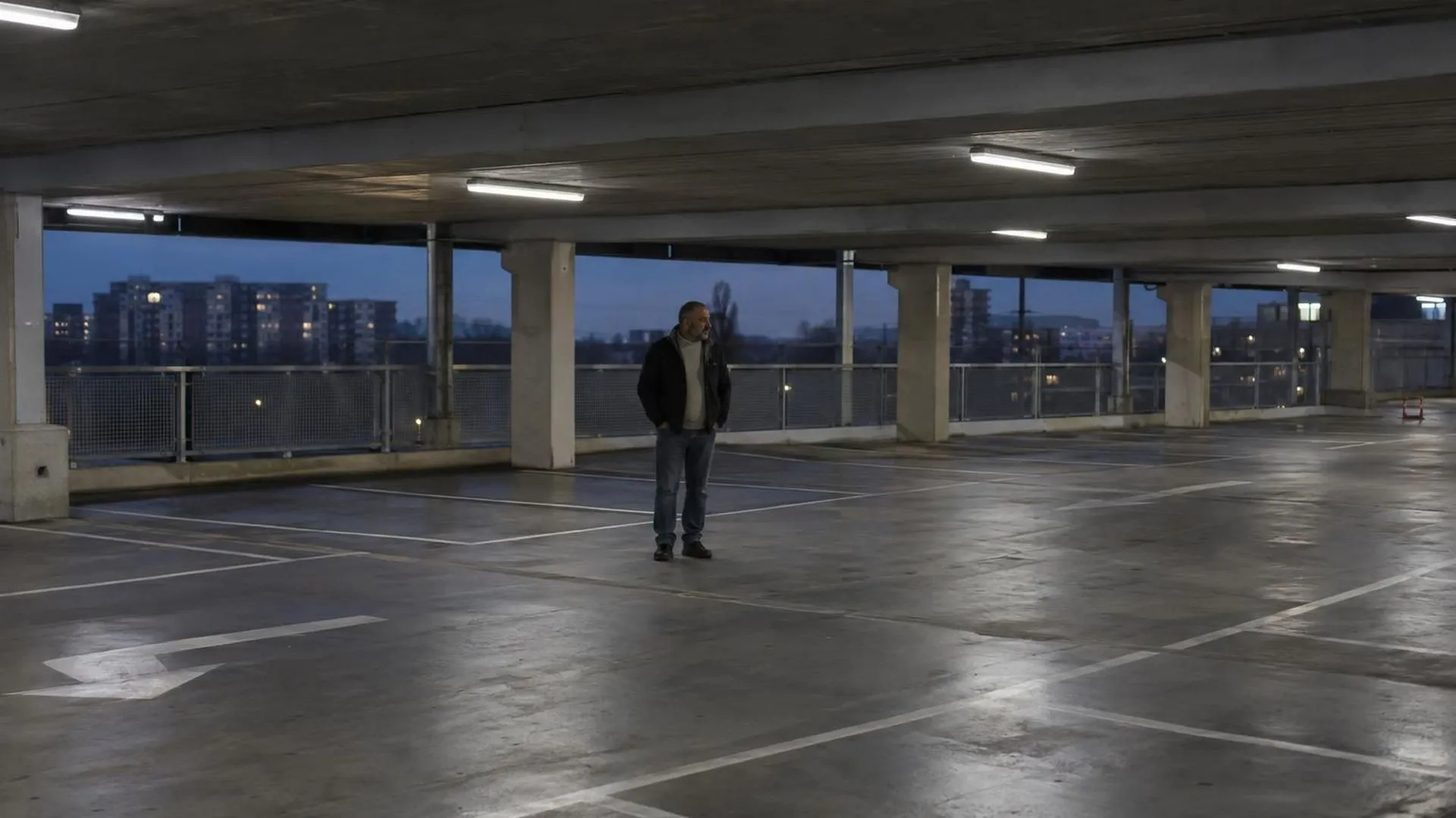 Iemand staat klein in een bijna lege parkeergarage onder fel kunstlicht.