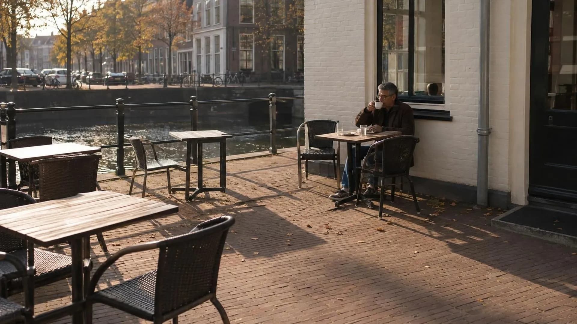 Op een bijna leeg terras in de vroege herfst zit iemand klein in beeld aan een tafeltje, met lange schaduwen van de laagstaande zon.
