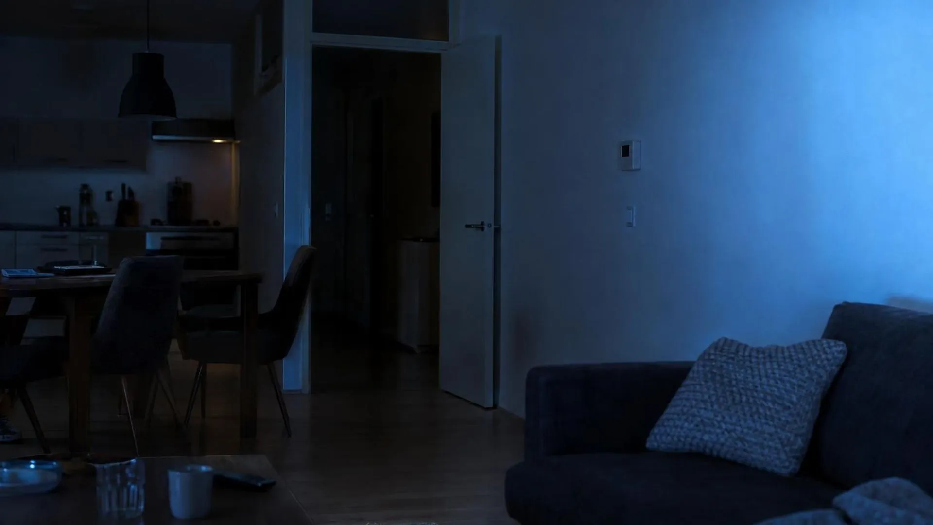 Het flikkerende blauwe licht van een tv weerkaatst tegen een woonkamerwand in een verder donker, stil appartement 's avonds.