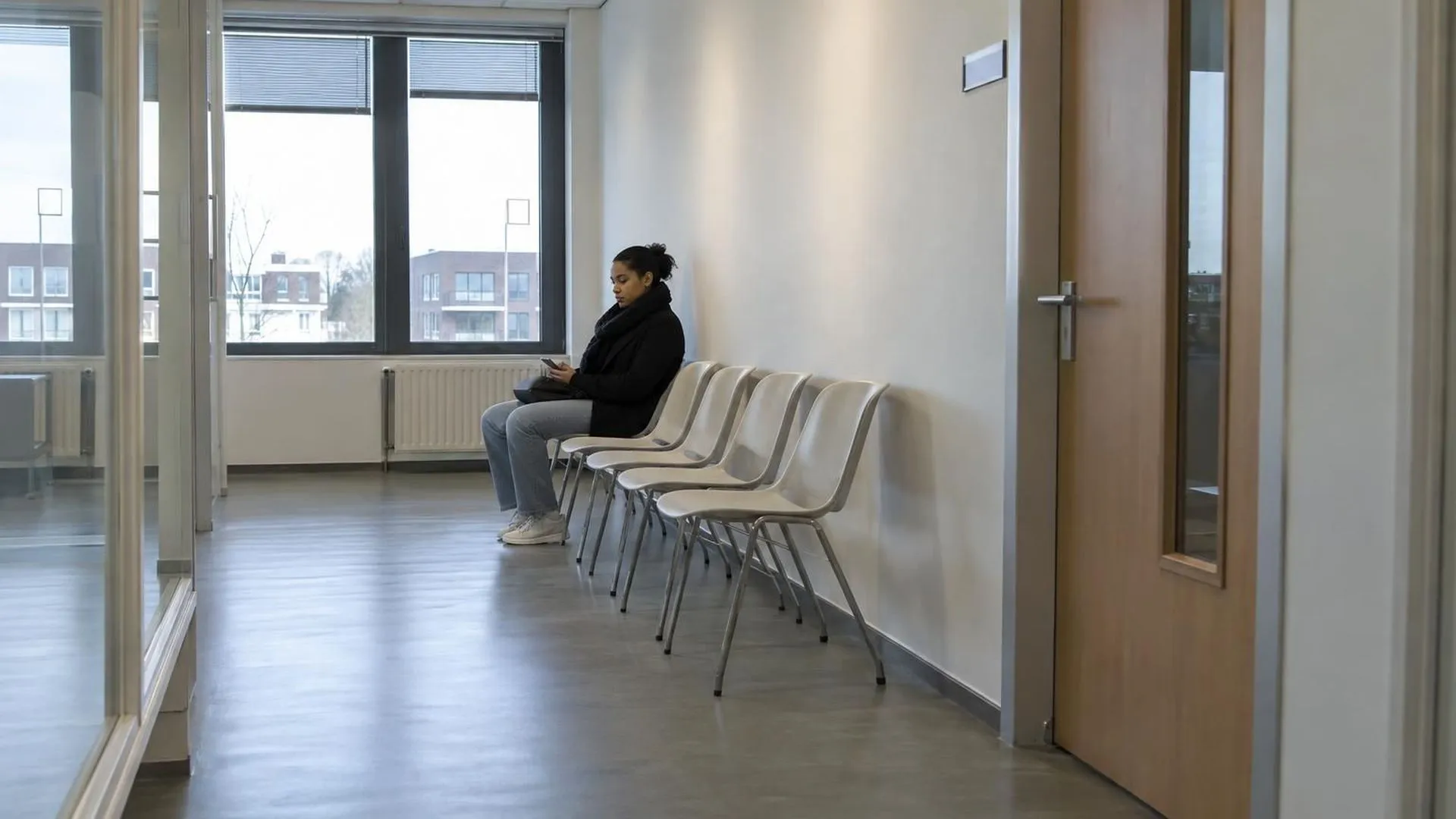 Iemand zit alleen op een rij plastic stoelen aan het einde van een lange, lege bedrijfsgang, wachtend voor een gesloten deur