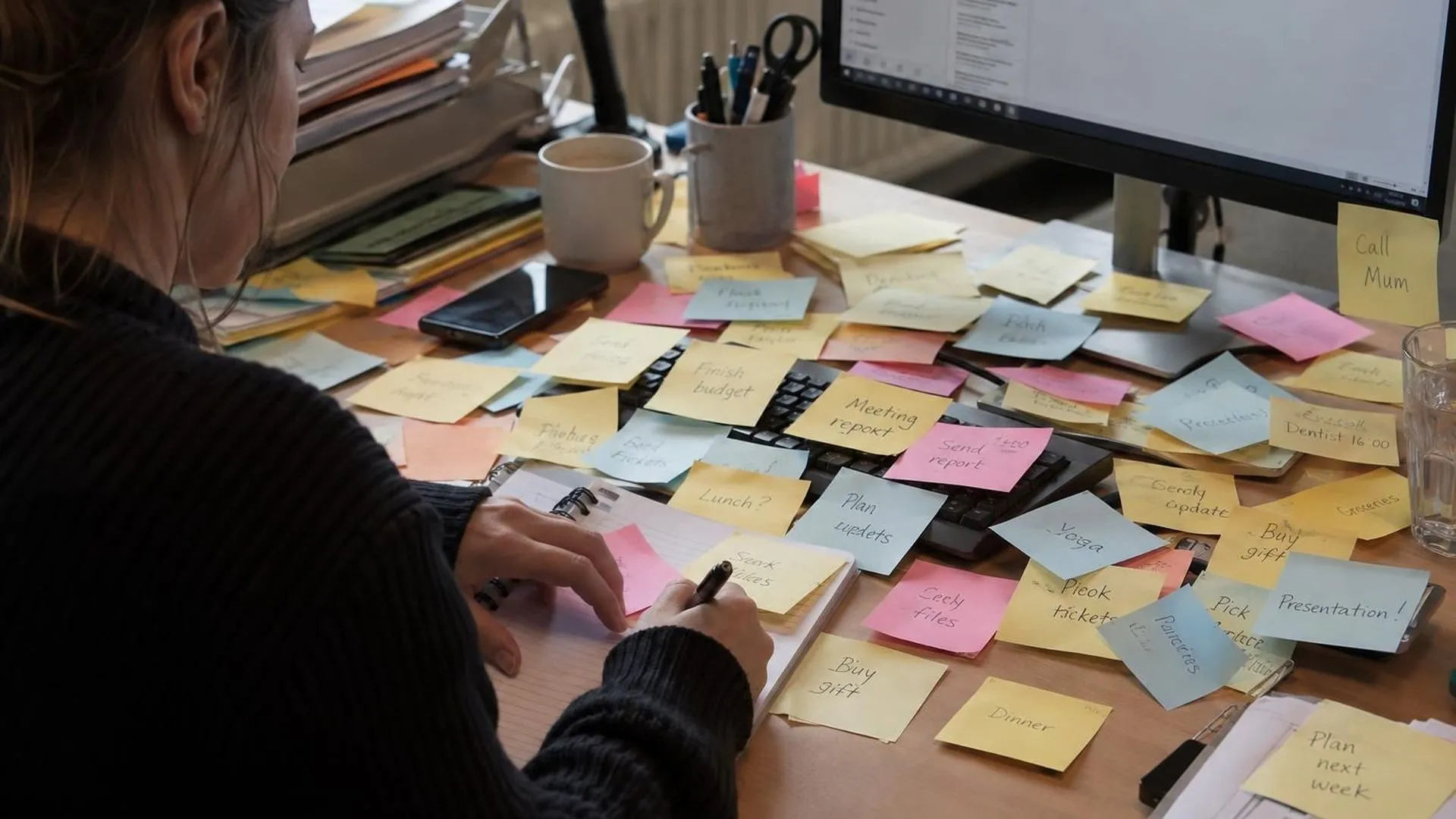 Een vol bureau met overlappende gele briefjes met steeds nieuwe taken erop, het toetsenbord er nauwelijks nog onder zichtbaar.
