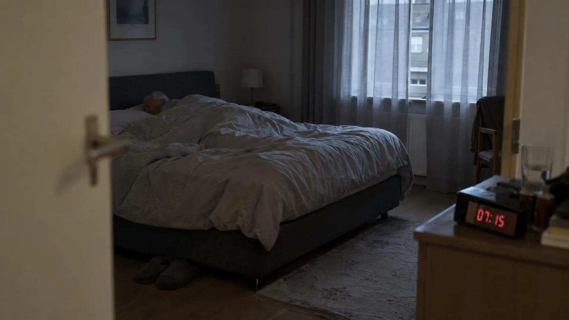 Een slaapkamer in grijs ochtendlicht, gezien vanuit de deuropening, met iemand nog onder de dekens.