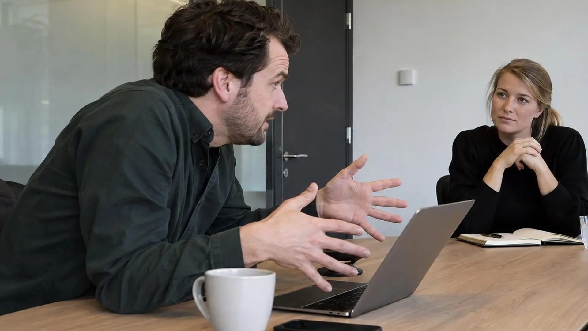 Twee mensen aan een tafel, de een leunt ver naar voren en neemt de ruimte in, de ander zit achterover met gevouwen handen
