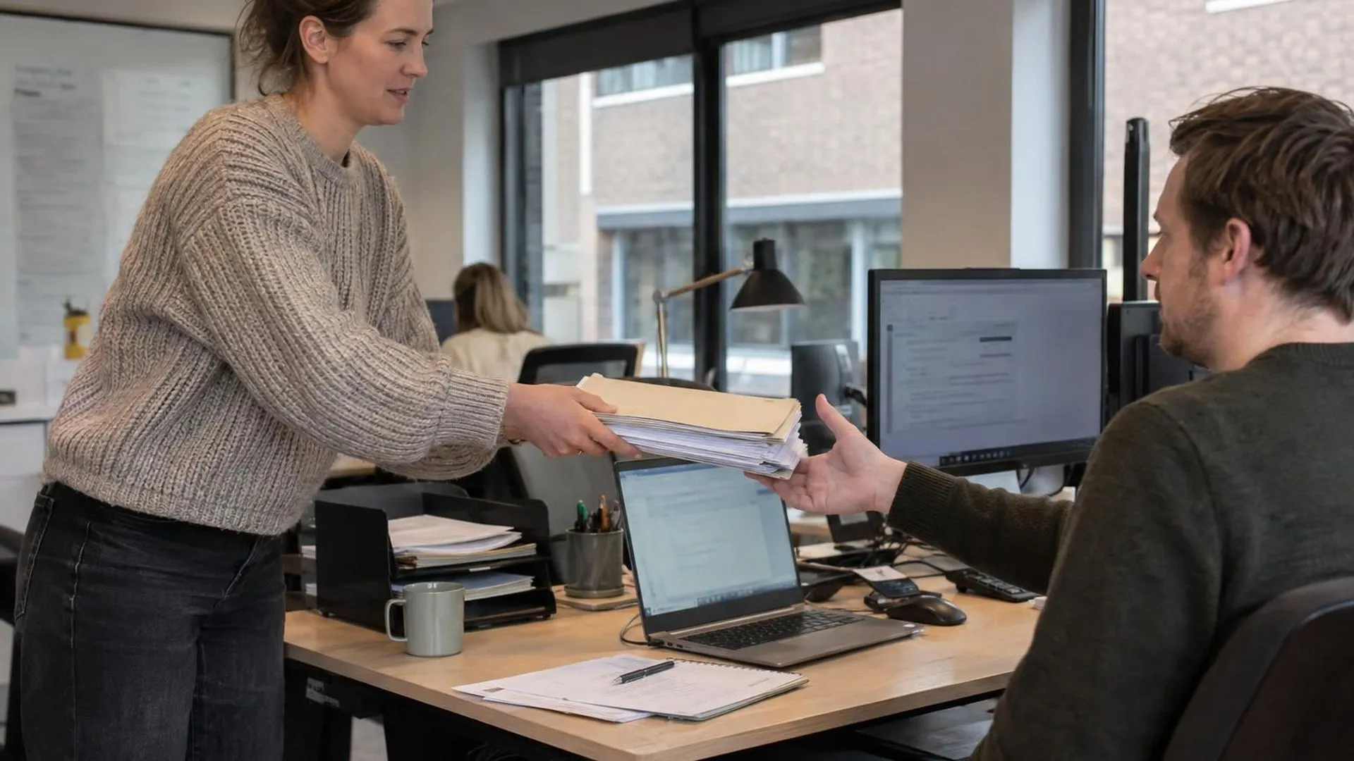 Een collega geeft een stapel dossiers aan een ander door, wiens hand al reikt om ze aan te nemen