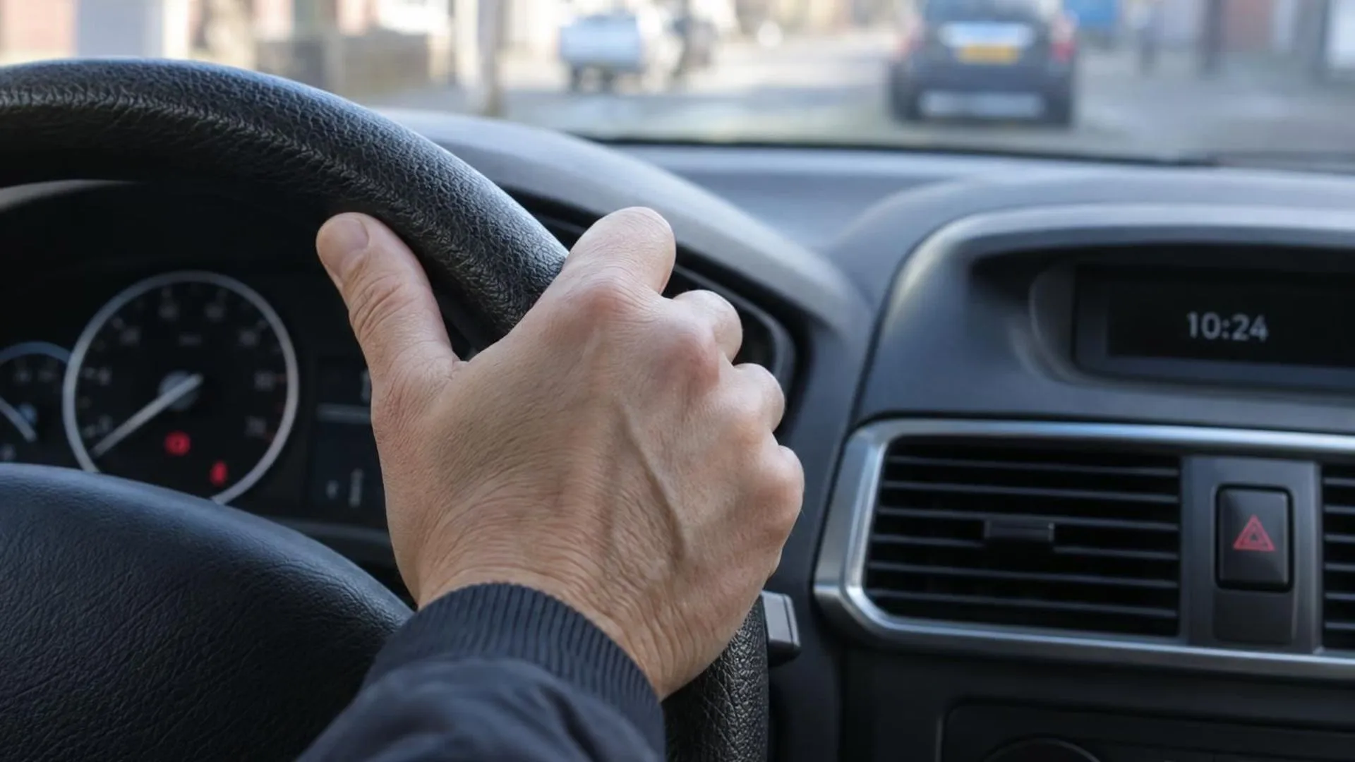Close-up van een hand die stevig het stuur van een geparkeerde auto vastgrijpt, met de klok op het dashboard zichtbaar