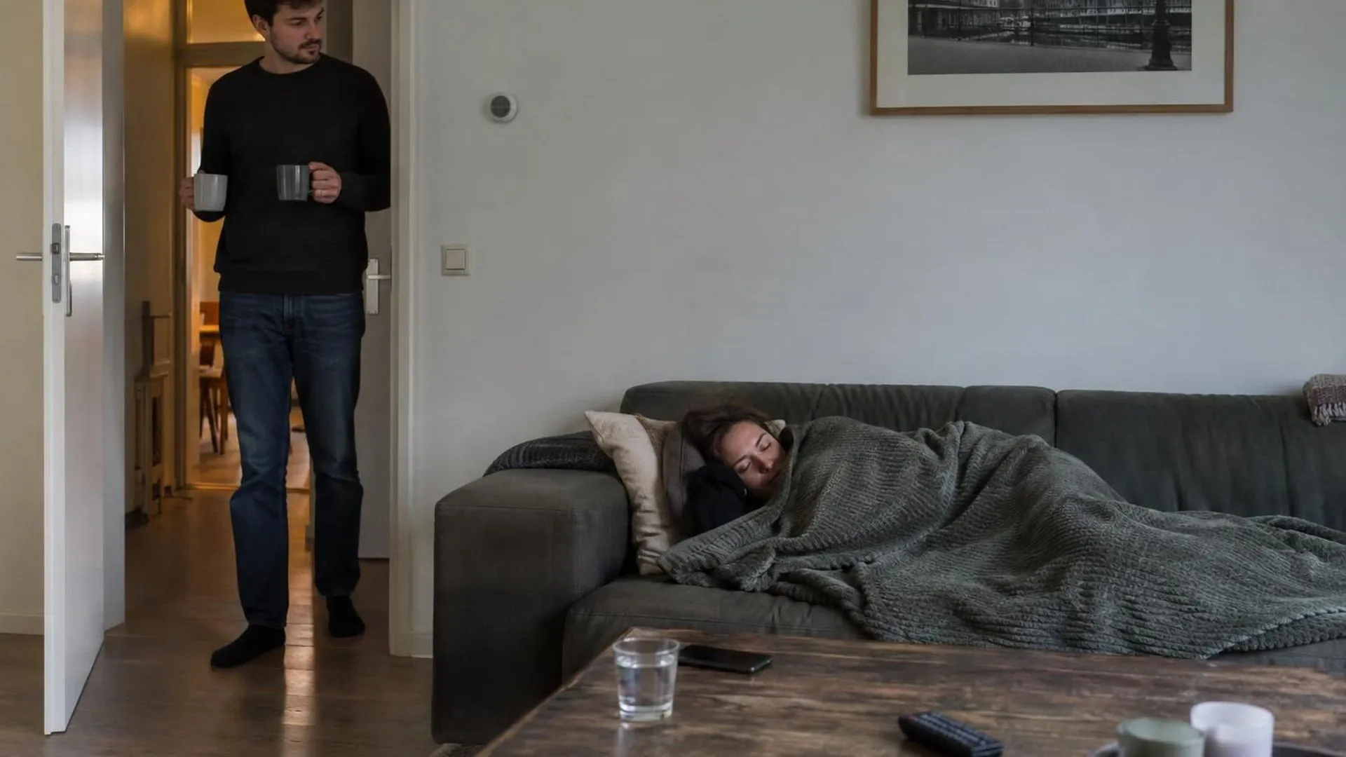 Twee mensen in de huiskamer: één ligt onder een deken op de bank, de ander aarzelt in de deuropening met twee mokken
