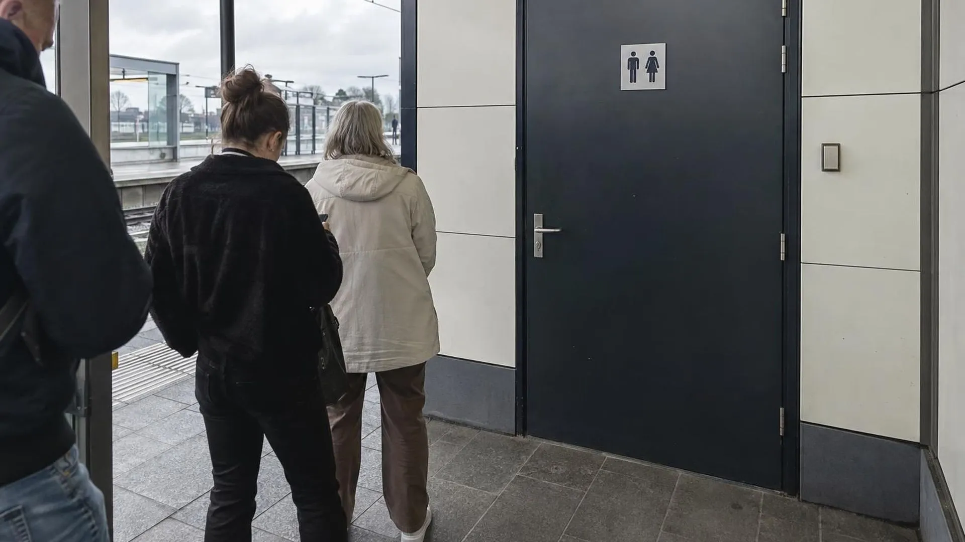 Een gesloten toiletdeur op een treinperron met een wachtrij ernaast.