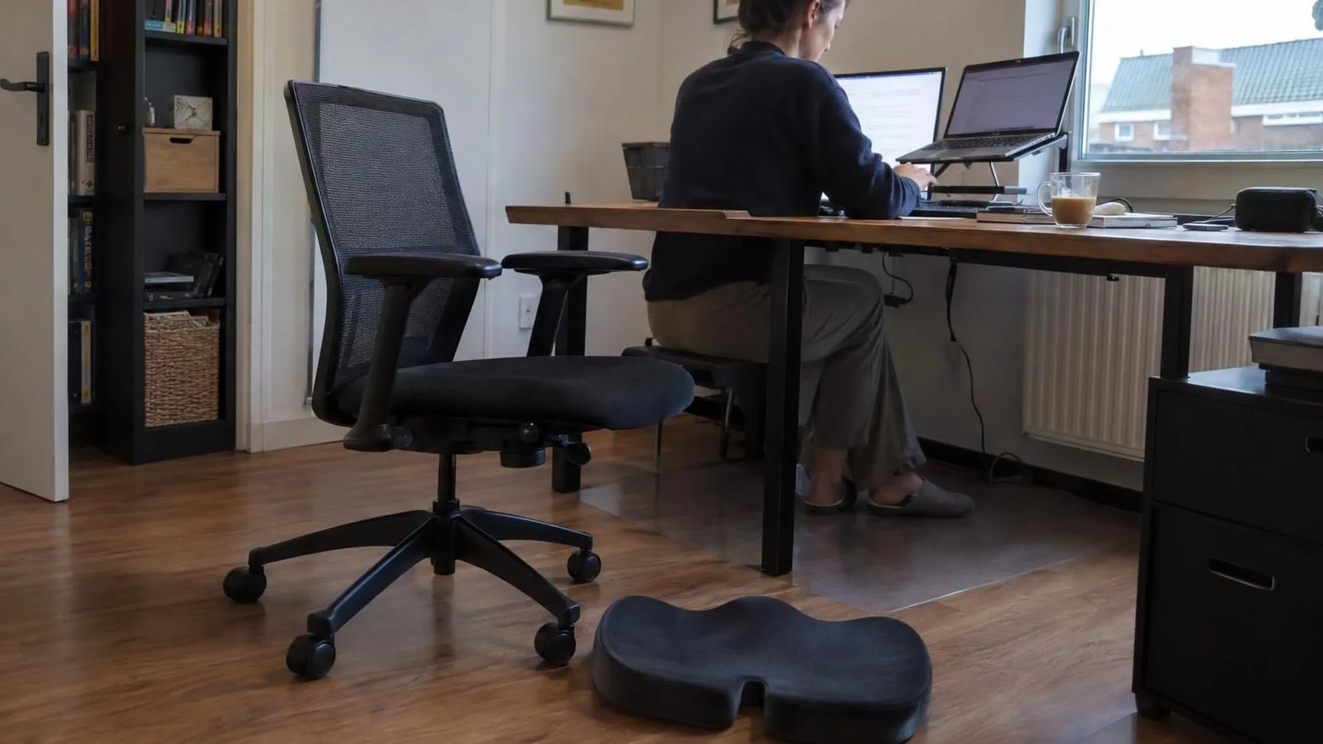 Een thuiswerkplek met een ergonomisch kussen dat op de grond ligt naast een lege stoel, laptopstandaard en koude koffie op het bureau