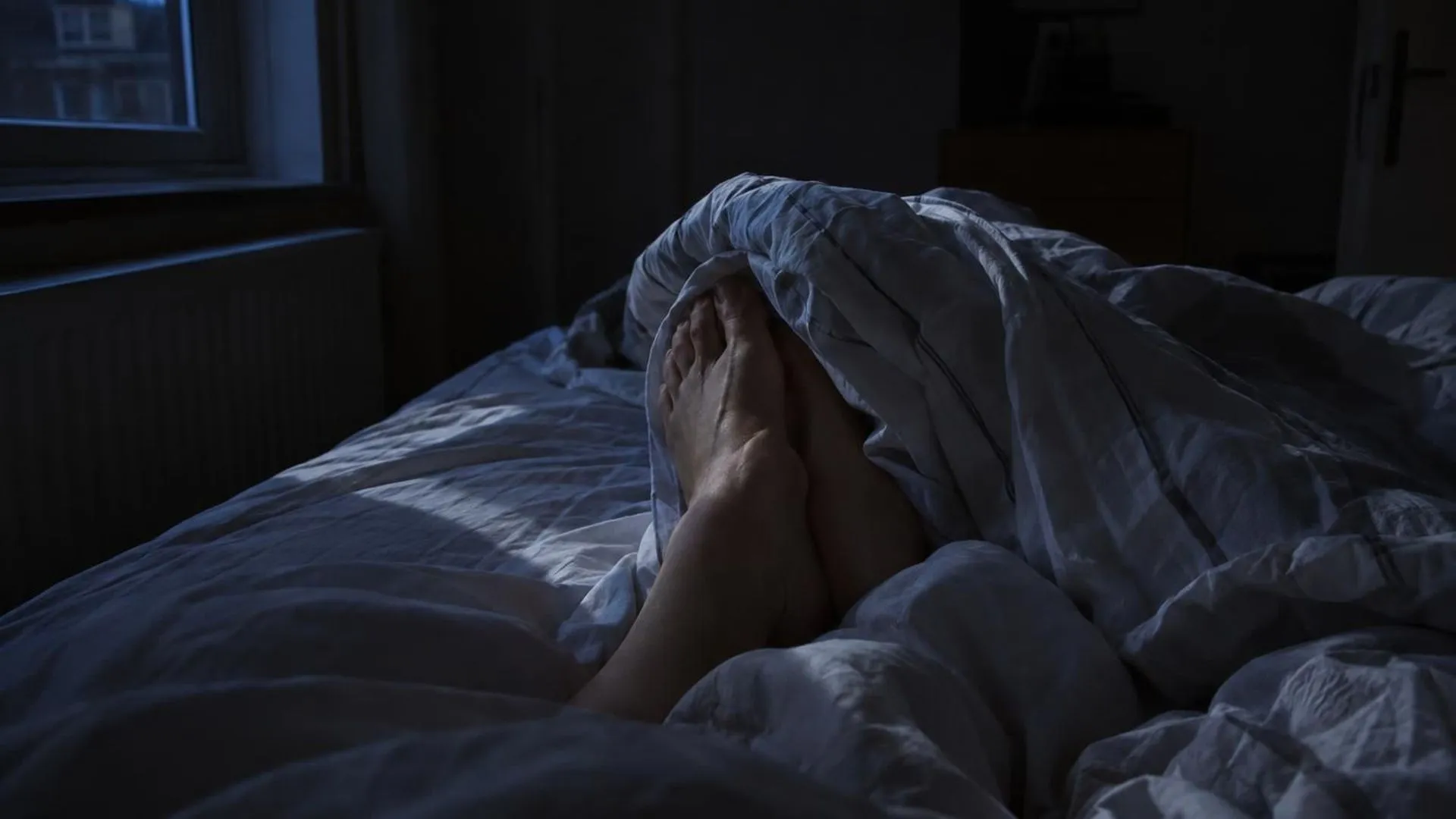 Close-up van blote voeten verstrikt in verwrongen lakens in het donker, met een streep maanlicht