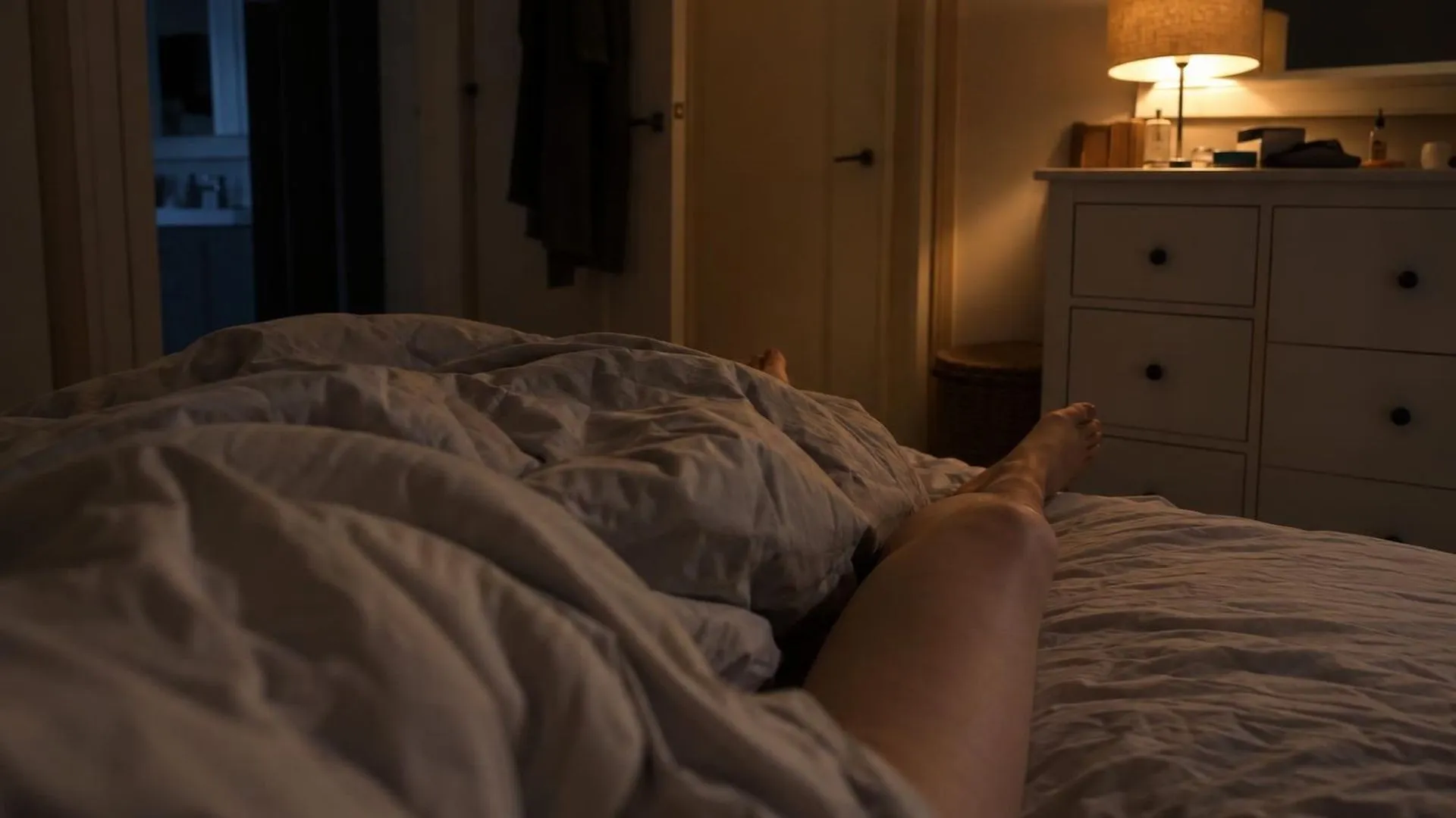 Close-up van een verkreukeld dekbed met benen die er half onderuit steken, in het gedempte licht van een nachtlampje.