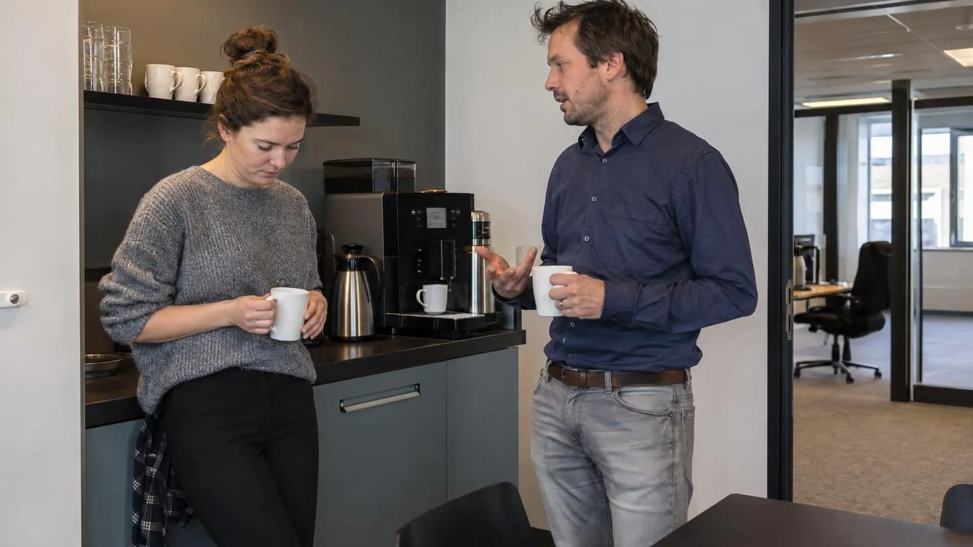 Twee collega's bij het koffieapparaat, waarbij een van de twee naar de grond kijkt terwijl de ander praat.